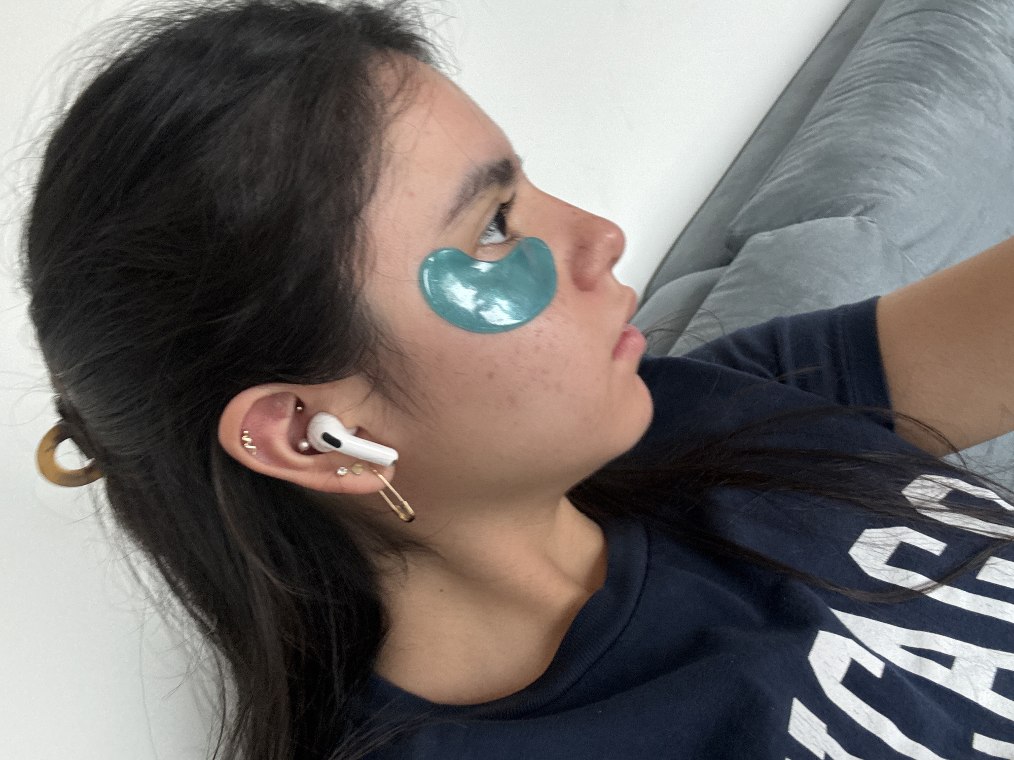 A woman is lying on a light blue couch, wearing a navy blue t-shirt with white lettering. She has dark hair, some pulled back with a brown hair clip. She is wearing an AirPod Pro in her left ear, along with multiple earrings, including a gold safety pin. A teal eye mask is placed under her left eye. The background consists of a white wall and the light blue couch. There is no information to determine the specific location in Austin, United States of America.