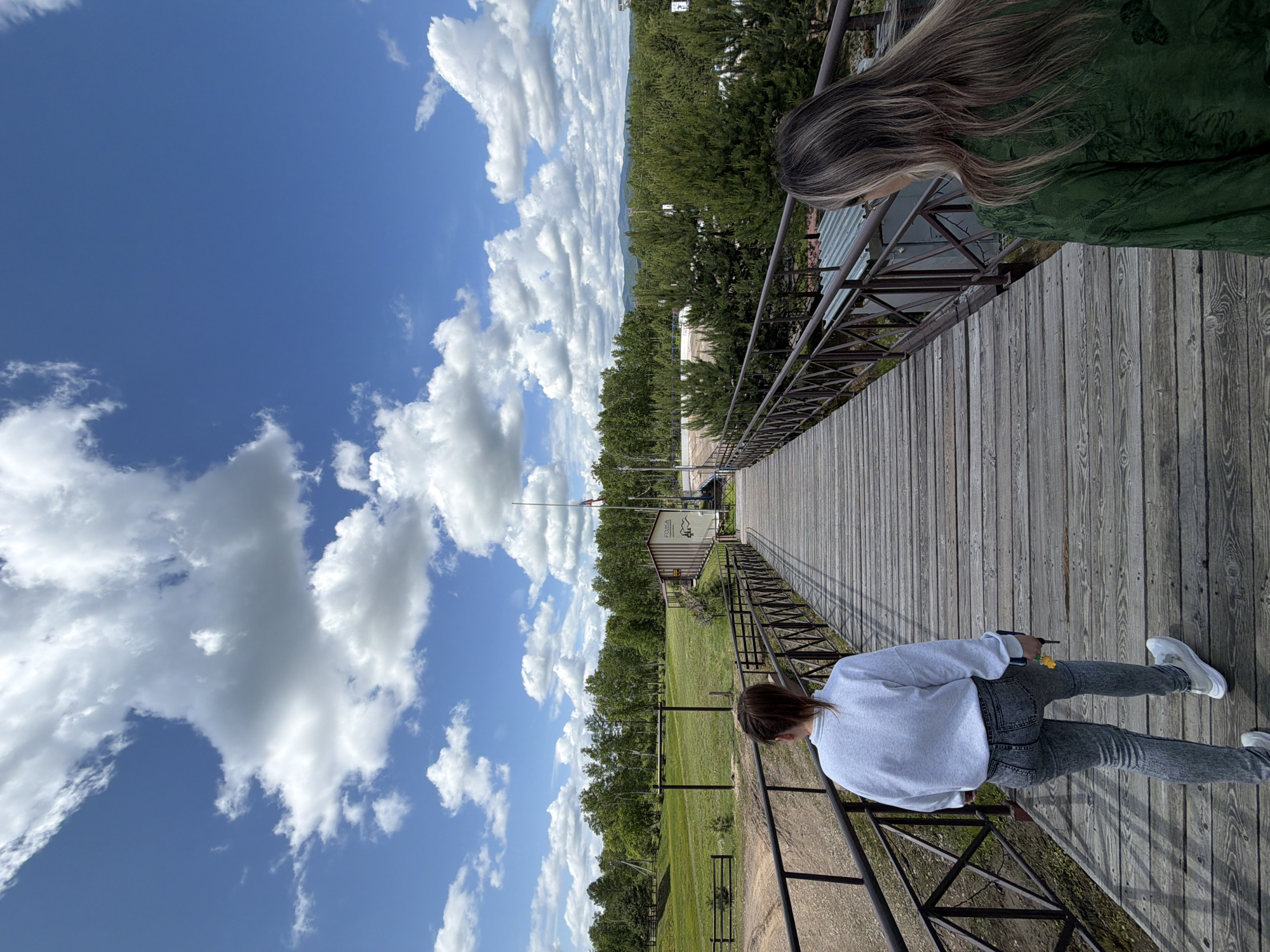 Two women are walking away from the camera on a wooden bridge. The woman on the left is wearing a white sweatshirt and grey jeans. The woman on the right is wearing a green patterned shirt. The bridge has dark brown metal railings. In the background, there is a green grassy field, a line of trees, and a building with a flagpole. The sky is blue with white, fluffy clouds. The image appears to have been taken outdoors on a sunny day. The location is the city of Birobidzhan, Russia.