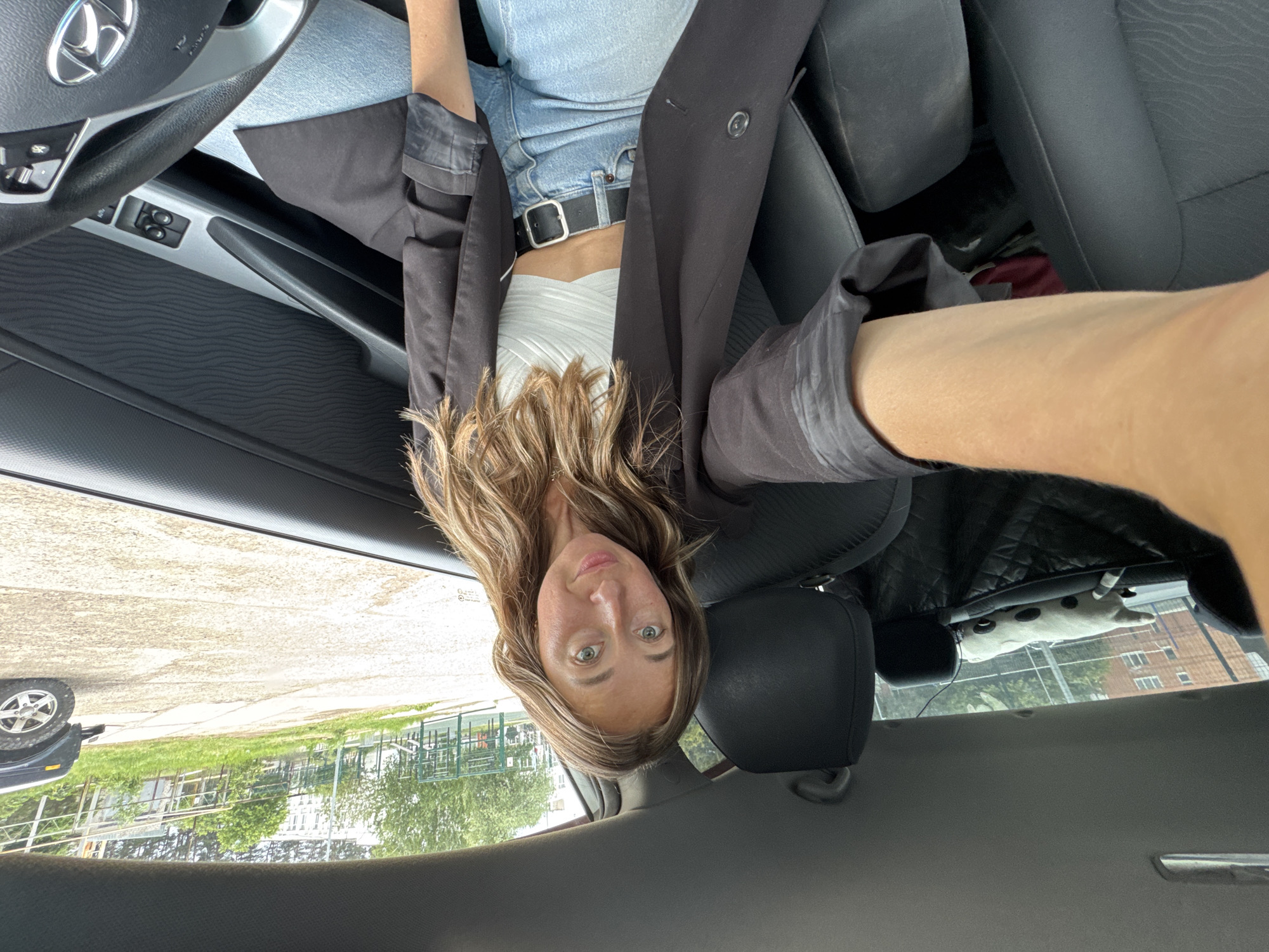 A head-on selfie captures a young woman seated in the driver's seat of a Hyundai car, looking directly at the camera with a neutral expression. Her long, wavy, light brown hair frames her face, revealing fair skin and light-colored eyes. She is dressed in a white ribbed crop top, light blue denim garment (likely jeans or shorts), and a black or dark grey blazer, cinched at the waist with a black belt featuring a silver buckle.
The car's interior is prominent, showing the black steering wheel with the Hyundai logo, the dashboard, and parts of the black door panel. The upholstery of the car seats is also dark-colored. In the back window, a white plush toy with black spots, possibly resembling a dog or cow, is visible on the parcel shelf.
Through the car windows, a daytime outdoor scene is visible. There is a paved area, green grass, and trees, with buildings (appearing to be multi-story structures) in the distant background. The wheel of another vehicle, possibly an SUV or jeep, can be seen partially through the driver's side window. The overall lighting suggests it is a bright day. A small, blurry sticker with text, possibly "DOT", is faintly discernible on the rear window. The setting is consistent with being in Chaikovsky, Russia.