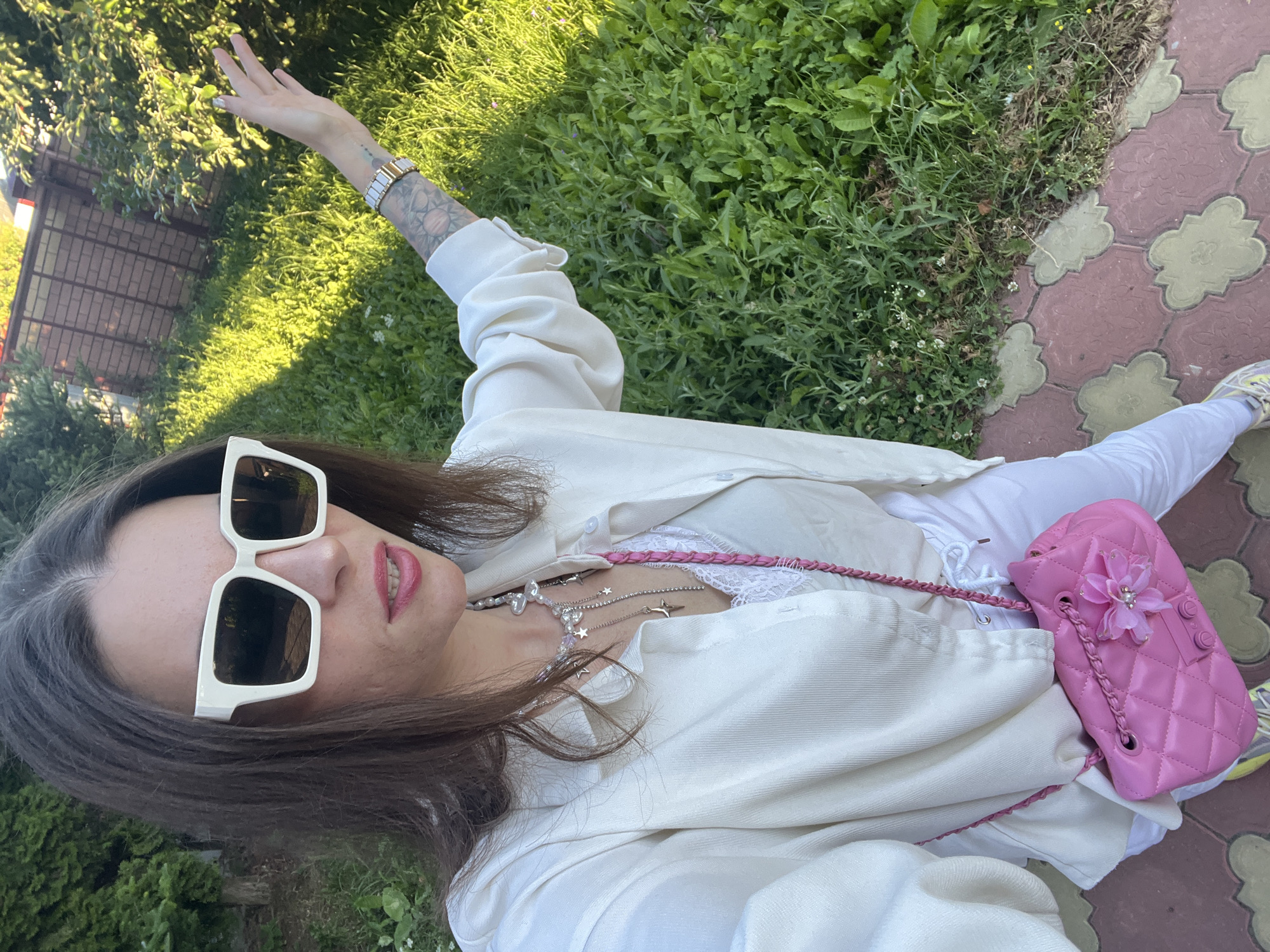 A woman is pictured outdoors in what appears to be a grassy area next to a paved walkway. The woman is wearing white clothing, including a button-down shirt and pants. She has on large, white-framed sunglasses and multiple necklaces, one featuring stars and pearls. A pink quilted handbag with a floral embellishment is slung across her chest. Her left arm is raised, and her hand is open. Tattoos are visible on her arm. The lighting suggests it is daytime, with sunlight casting shadows. The scene is likely a park or garden given the presence of grass and trees. The woman's expression is difficult to discern due to the sunglasses and the angle of the photo. There is no visible text in the image.