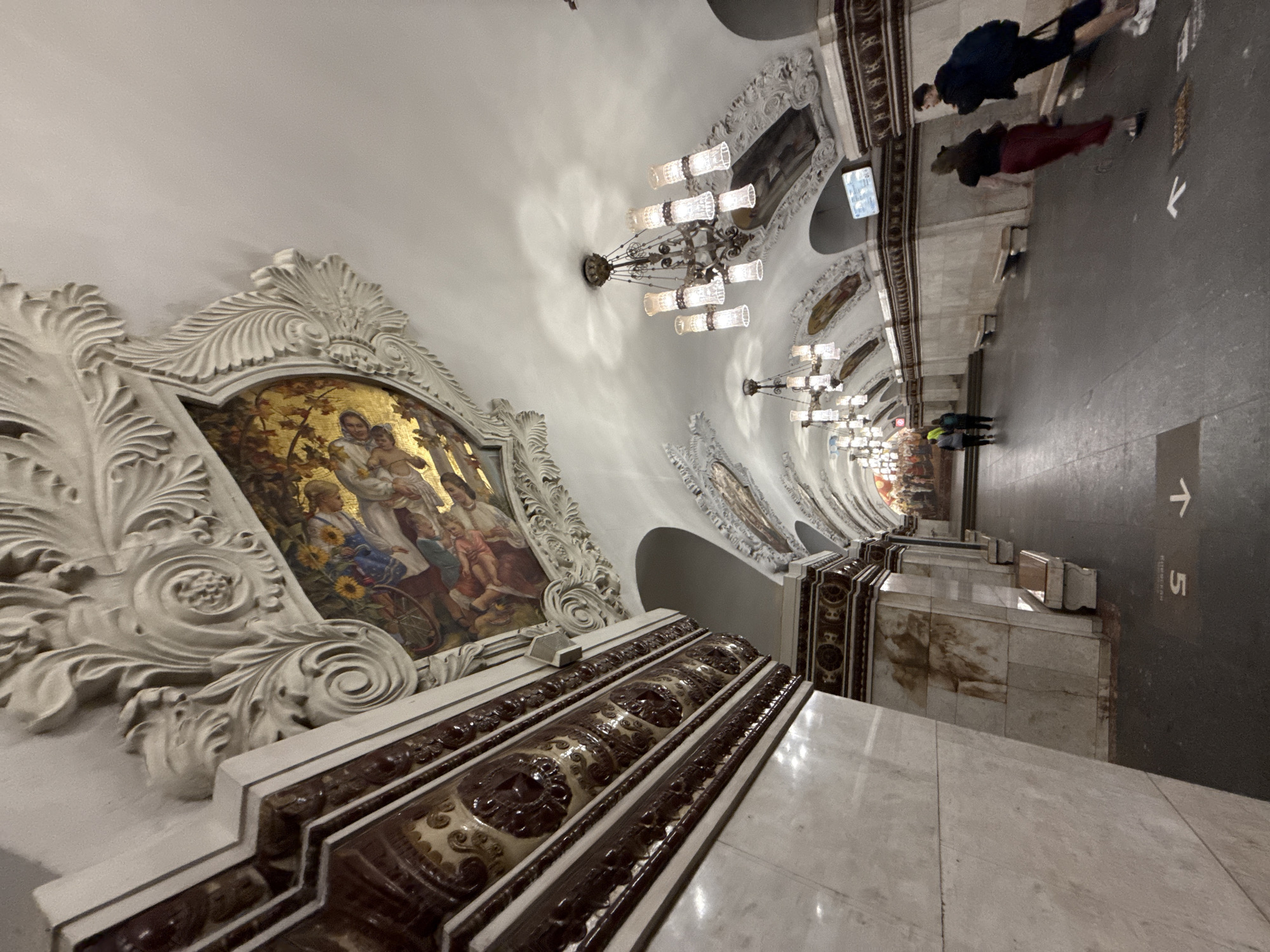 The image depicts a view within a Moscow Metro station, characterized by opulent architectural details. The ceiling features elaborate stucco work and ornate murals depicting scenes with figures and floral motifs, accented with gold leaf. Multiple chandeliers with numerous glass tube-shaped lights hang from the ceiling, illuminating the vaulted passageway. The walls are clad in marble with decorative friezes featuring intricate patterns and dark, rich-colored tiles. Benches line the sides of the platform. Several individuals are visible walking along the platform. Two people, one wearing a dark top and red trousers, the other in a dark top and black shorts with a backpack, are in the foreground, walking away from the camera. Further down the platform, two individuals in darker clothing are walking away from the camera, and a few more people are seen in the distance. Arrows and numbers are marked on the floor, indicating direction and possibly platform numbers. The overall impression is one of grandeur and historical significance, typical of many Moscow Metro stations.