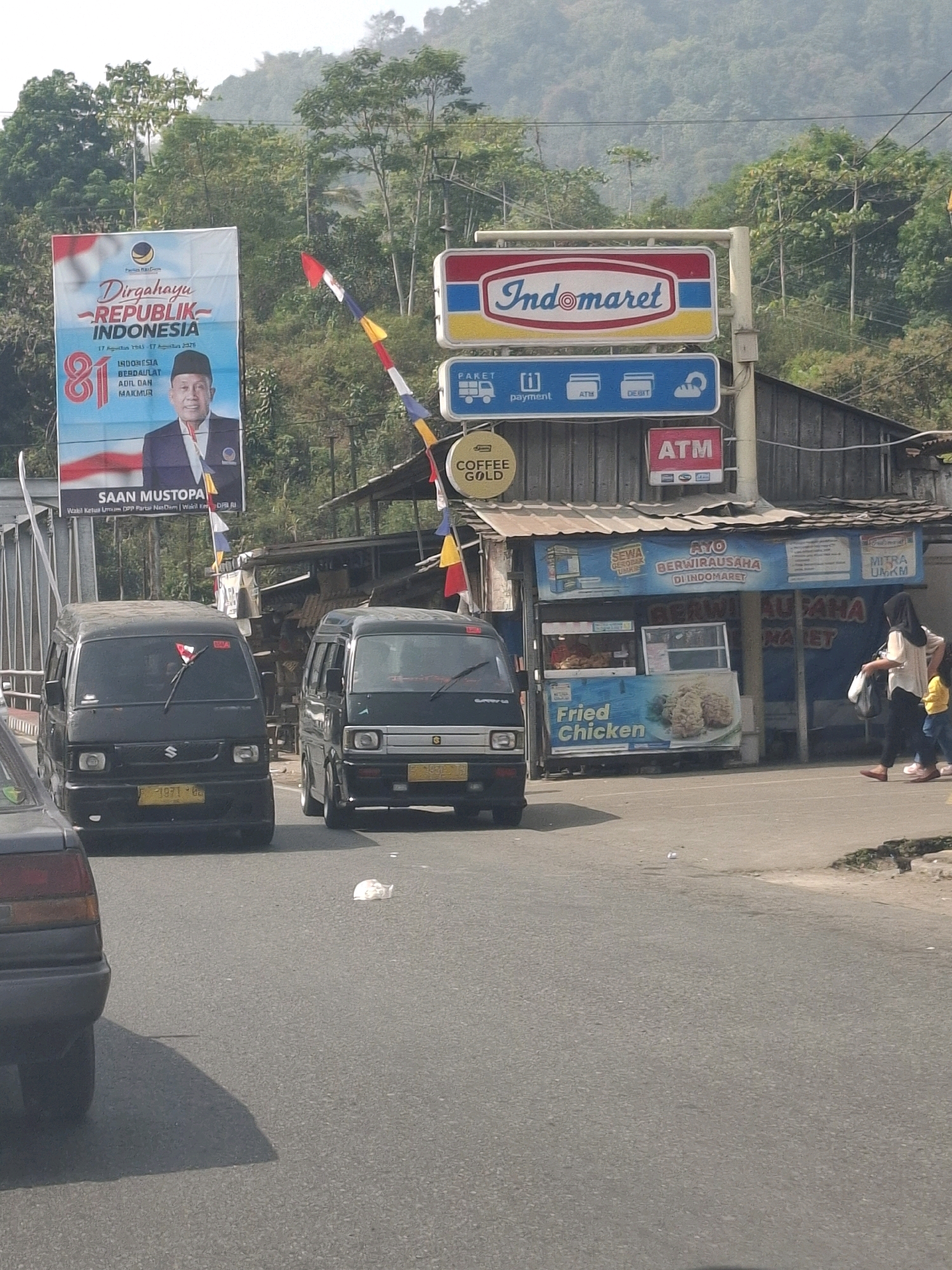 The image shows a street scene in Sasagaran, Indonesia, with a focus on a roadside Indomaret convenience store. Two black vans are parked on the road, one in the foreground with its license plate "F 1971 BG" partially visible. Behind the vans, the Indomaret store displays advertisements for "Fried Chicken" and "Coffee Gold." A banner celebrating "Dirgahayu REPUBLIK INDONESIA" with a portrait of Saan Mustopa is also visible. A woman in a hijab and a child are walking on the sidewalk in front of the store. The background features lush green hills and trees, indicating a somewhat rural or semi-urban setting. The sky is overcast, and the overall lighting suggests it is daytime.