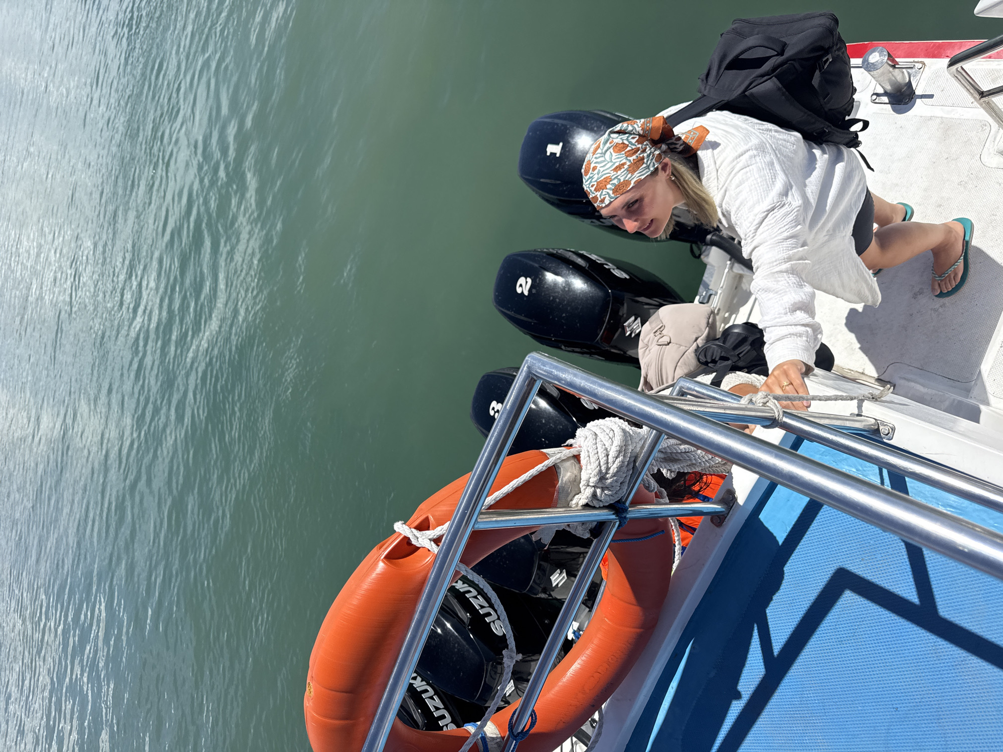 An overhead, slightly angled shot captures a young woman on the stern of a boat, situated in calm, greenish-blue water under a bright sky. The woman, with light-colored hair, wears a white long-sleeved cover-up, black shorts, and turquoise flip-flops. A vibrant orange and blue-green patterned headscarf covers her hair. She is seen in a leaning posture, holding onto a silver metal railing, with a black backpack resting on her back. Her gaze is directed downward and forward, towards the water.
Behind her, three black outboard boat engines are prominently visible. The brand name "SUZUKI" is identifiable on their sides, and the numbers "1," "2," and "3" are clearly displayed on their top covers. An orange lifebuoy, secured by ropes, is positioned near the railing towards the bottom left of the frame. The boat's deck features both white and blue non-slip surfaces.
The water appears tranquil with subtle ripples, reflecting the bright daylight. Strong, defined shadows of the boat's railing and structure are cast onto the blue deck, indicating a sunny day. The scene suggests a maritime activity or journey, likely taking place during the daytime in a warm climate like Denpasar, Indonesia.