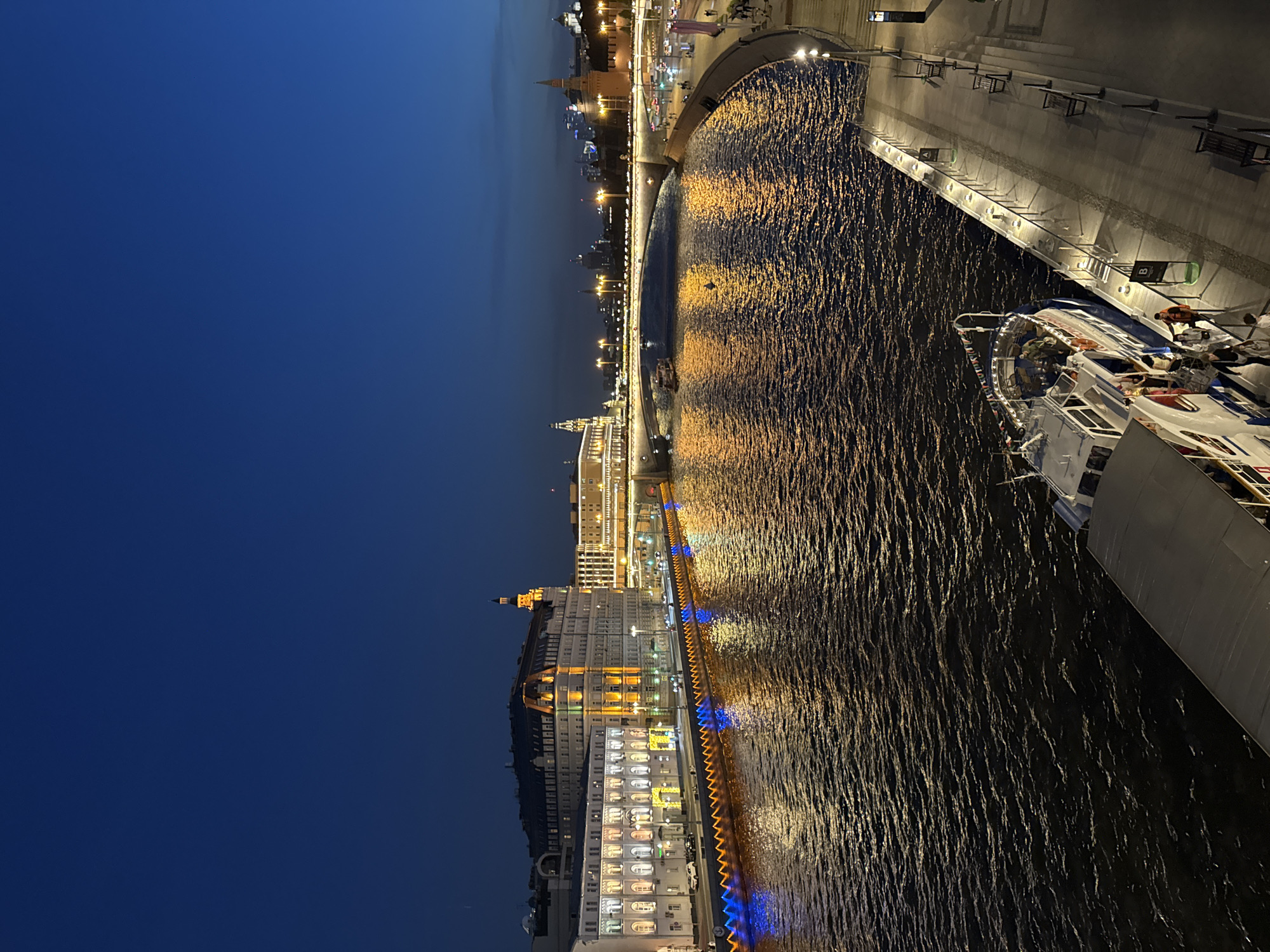 The image displays a nighttime panoramic view of the Moscow River in Moscow, Russia, looking east towards the city center. The dark blue sky transitions subtly to a lighter hue near the horizon.
On the left side of the frame, along the Sofiyskaya Embankment, a large, multi-story building, identifiable as the Baltschug Kempinski Hotel, is prominently illuminated with warm yellow and white lights, highlighting its architectural details and numerous windows. Adjacent to it are smaller, lighter-colored buildings. The embankment itself is adorned with a sequence of alternating blue and orange lights, which create vibrant reflections on the dark, rippling surface of the river.
In the mid-ground, the Bolshoy Kamenny Bridge (Large Stone Bridge) spans the Moscow River, illuminated along its arches and roadway. Beyond the bridge, on the far bank, several towers of the Moscow Kremlin and parts of its historic wall are discernible against the urban skyline, along with other illuminated buildings of the city center. A smaller, illuminated vessel is visible on the river near the bridge.
In the right foreground and mid-ground, a wide, paved pedestrian embankment features evenly spaced lampposts, several benches, and a railing. A modern boat dock extends into the river, marked with signs "A" and "B" and illuminated by ground-level lights. A large passenger boat, a river cruise vessel, is moored at the dock. Several individuals are visible on the dock and the boat, appearing to be boarding or disembarking. One person on the dock is wearing an orange life vest. Further down the embankment on the right, additional pedestrians are visible. The river water intensely reflects the golden and orange artificial lights from the surrounding structures.
