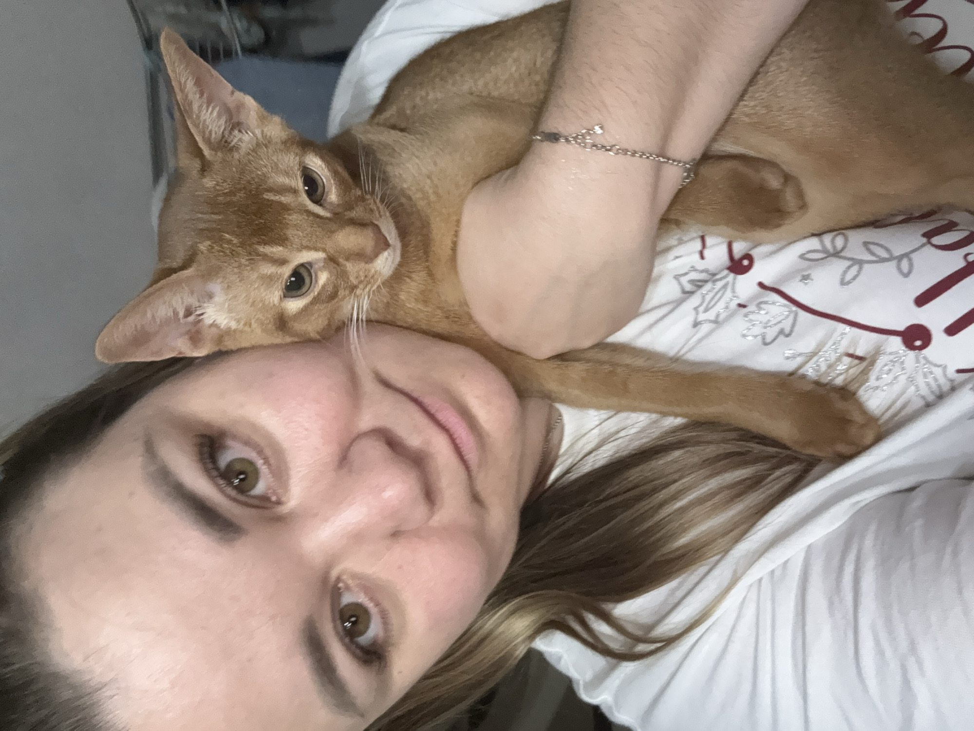 A close-up selfie shows a Caucasian woman holding an Abyssinian cat. The woman has brown eyes, fair skin, and blonde hair pulled back. She is wearing a white t-shirt. The cat is reddish-brown with large ears and is looking down. The background is blurred and appears to be indoors. The image is a selfie taken by the woman. There is no information that can be used to determine the location in Stary Oskol, Russia, or any other specific location.