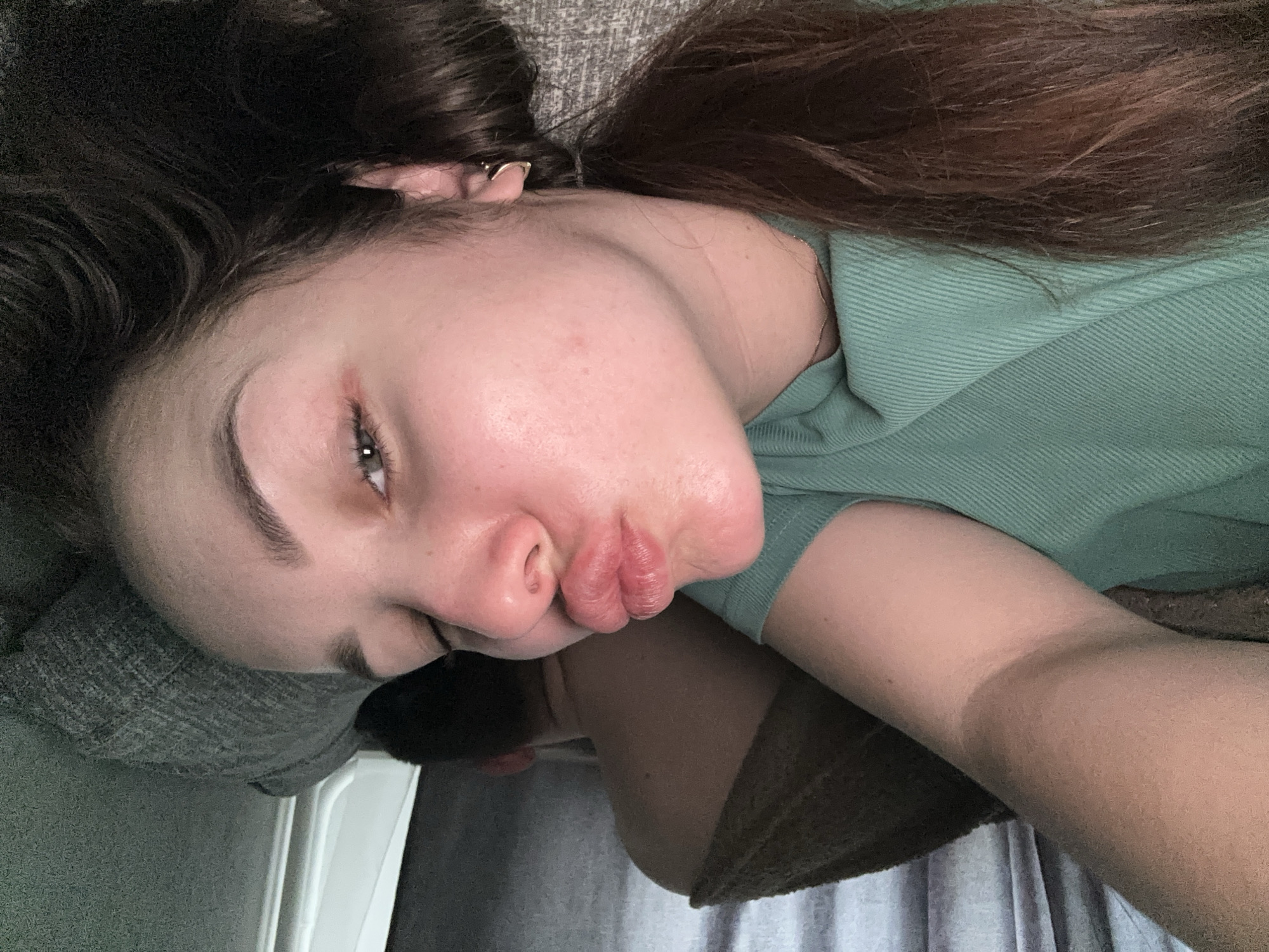 A young woman with long, brown hair and fair skin is lying on what appears to be a gray sofa or bed. She is wearing a light green shirt. Her face is tilted towards the camera, and her lips are pursed. An ear piercing is visible in her left ear. The background is blurred and shows fabric of various textures and colors. The location is not discernible from the image.