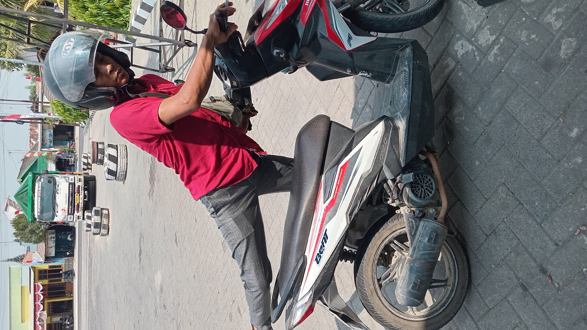 A person wearing a red shirt and gray pants, with a black helmet, is posing on a motorcycle. The person's right leg is extended backwards, and their left hand is on the motorcycle's handlebar. The motorcycle is black and red with "Beat" written on the side. The scene appears to be in an outdoor paved area, possibly a parking lot, with buildings and a truck in the background. There are stacked tires visible in the background, as well as some foliage. The lighting suggests it is daytime.