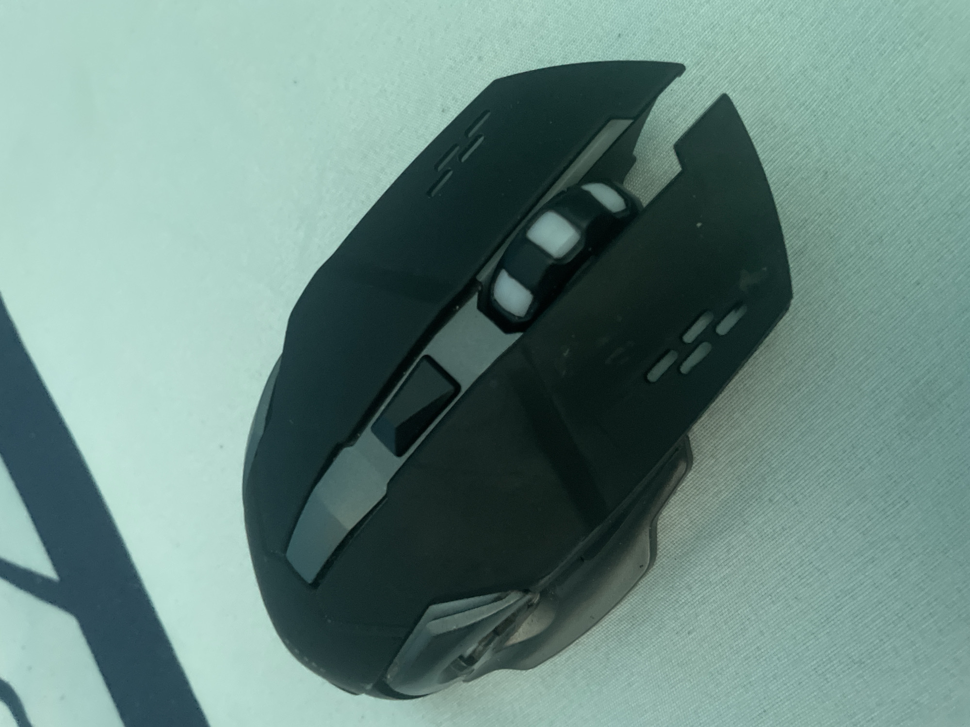 A black gaming mouse with a scroll wheel is positioned on a light blue textured surface, likely a cloth. The mouse features a sleek, angular design with visible buttons and what appears to be a clear plastic accent on the left side. The image does not provide any visual cues or context to suggest a location in Kołobrzeg, Poland.