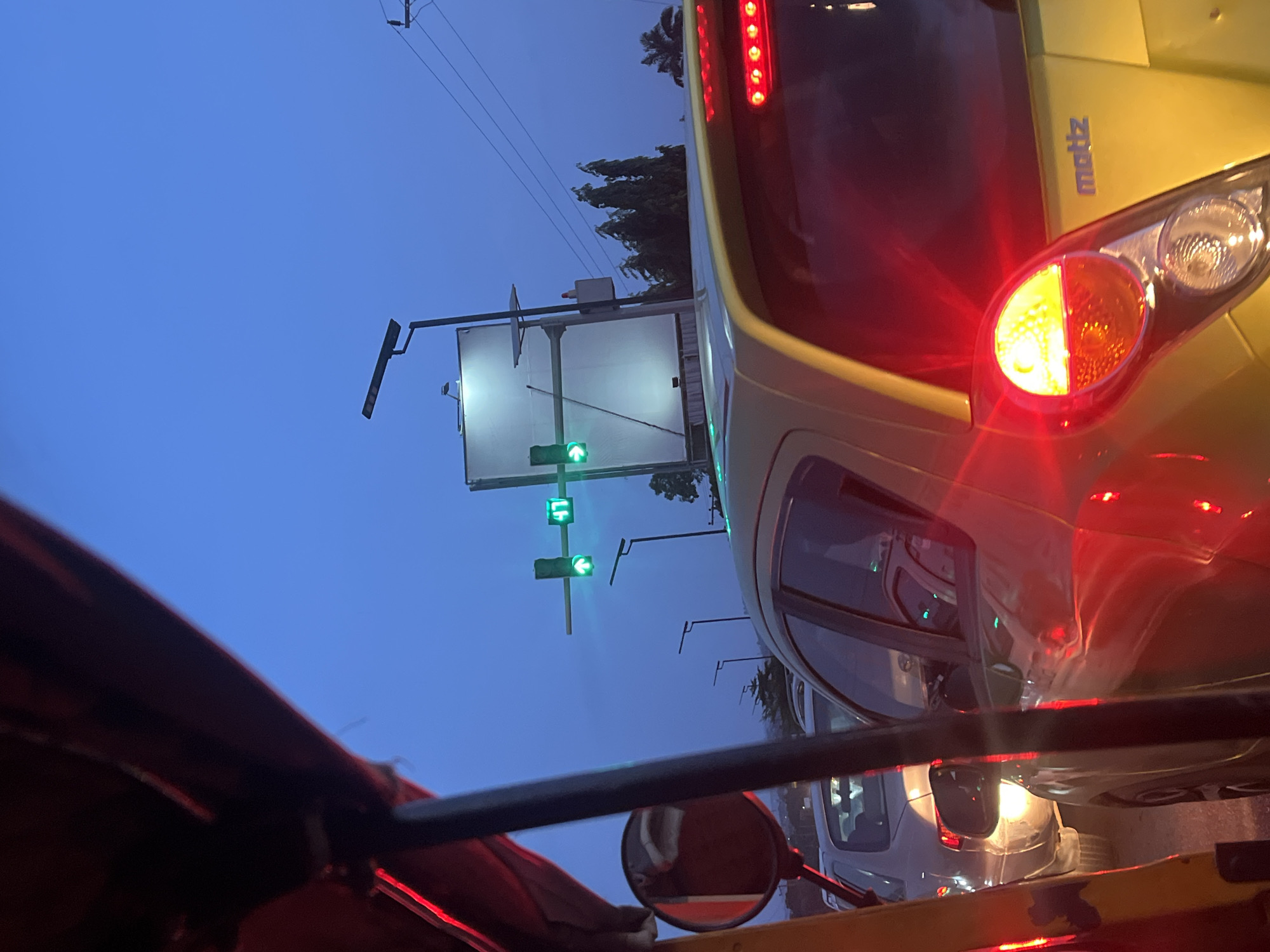 The image depicts a street scene in Galadimawa, Nigeria at dusk. The view is from inside a vehicle, likely a taxi or auto-rickshaw, looking out at traffic. A yellow "matiz"-branded vehicle is prominent, with its right turn signal activated. A traffic light shows a green arrow. Other vehicles are visible in the background. The sky is a deep blue. A street sign or advertisement board is also visible.
