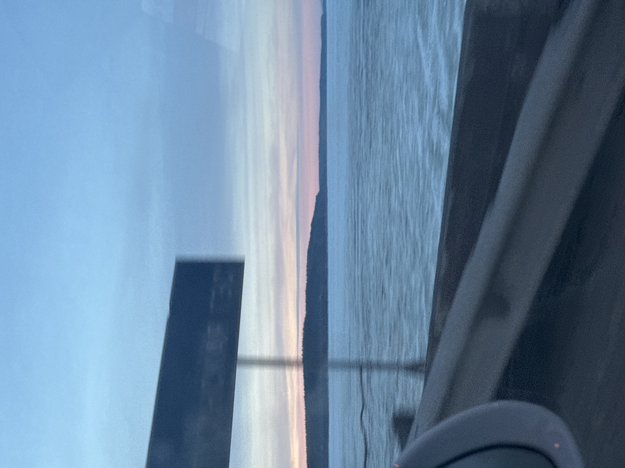 The image captures a scene from a moving vehicle, providing a view of the Votkinsk Reservoir in Chaikovsky, Russia, during twilight. In the immediate foreground, a dark gray vehicle side mirror is partially visible on the lower left, and a dark, horizontal guardrail or bridge barrier extends across the bottom. A large, dark rectangular sign, heavily motion-blurred and out of focus, occupies the upper-left quadrant, obscuring specific text but indicating an overhead road sign. This sign is supported by a thin, dark vertical pole extending downwards. The expansive mid-ground displays the Votkinsk Reservoir, its surface appearing light blue and textured, consistent with either partially frozen water or a choppy, icy river. In the background, a dark, undulating landmass, seemingly forested, stretches along the horizon line. The sky above transitions from a light blue at the zenith to subtle bands of light blue, pink, and orange closer to the horizon, indicative of either sunrise or sunset. No people are discernible in the frame.