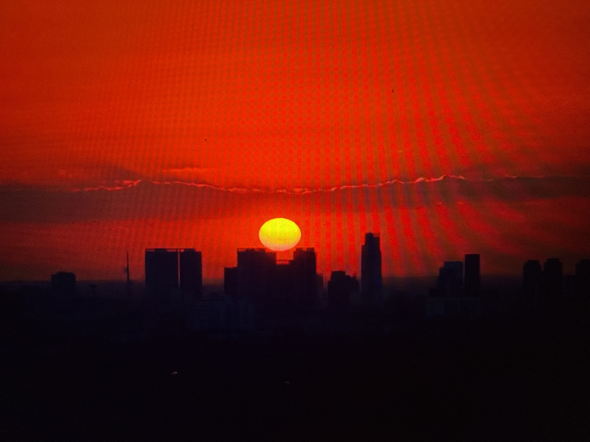 The image depicts a city skyline silhouetted against a vibrant red sky. A large, bright yellow sun is partially visible behind the buildings, creating a dramatic sunset effect. The sky is dominated by intense red hues, with vertical lines of light suggesting either a digital display or a specific atmospheric phenomenon. The city's buildings are dark and indistinct, their shapes defined only by their outlines against the fiery sky. The foreground is a dark, shadowy expanse, obscuring any ground-level details. There are no people or specific objects clearly identifiable within the scene.