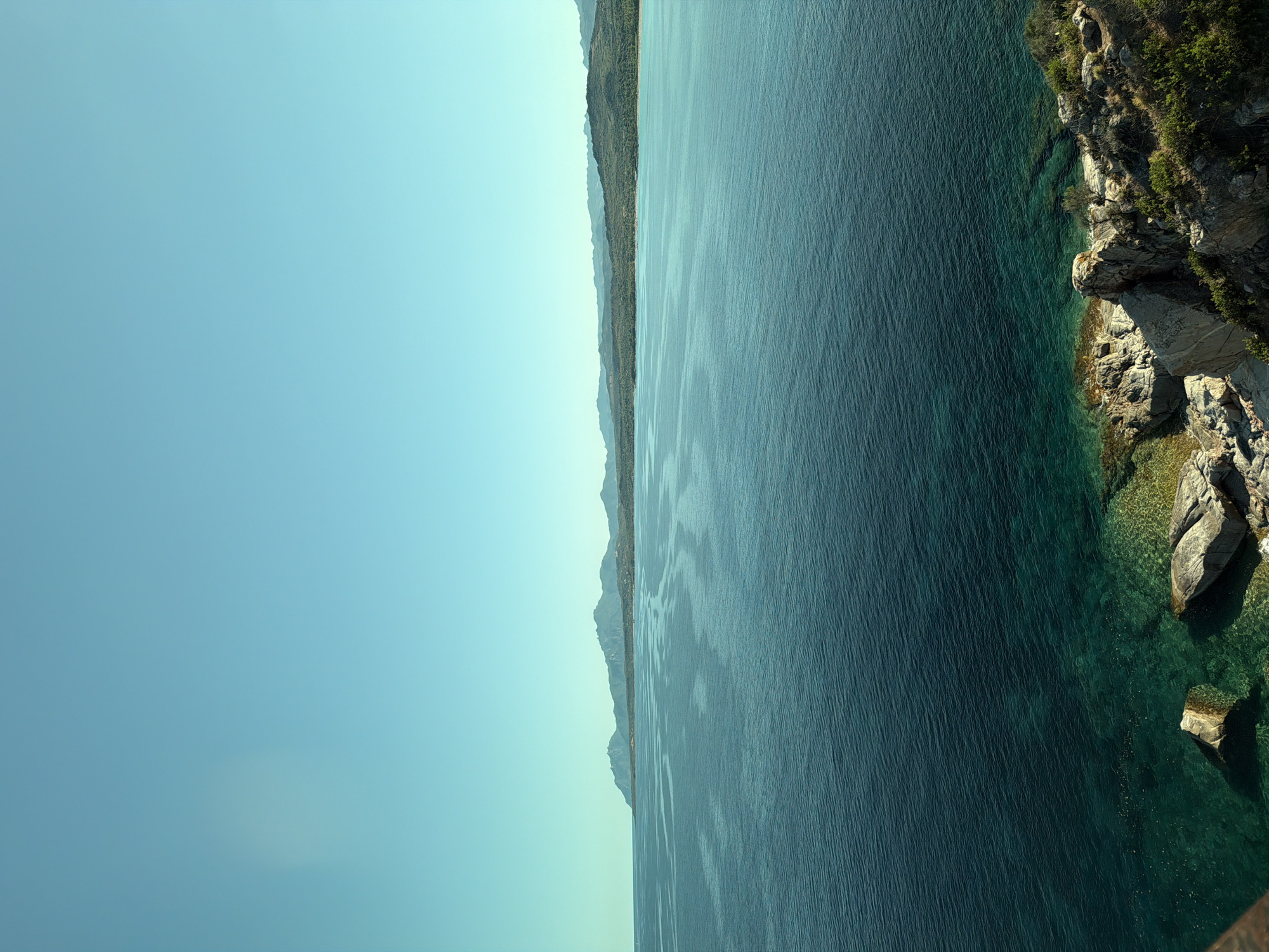 The image depicts a scenic coastal view of the sea and the distant coastline, likely near Tortolì, Italy. The sky is a clear, pale blue, with a faint, circular light artifact on the left side, possibly from the camera lens. The sea occupies the majority of the frame, transitioning from a deep, dark blue in the foreground to a lighter, more turquoise hue closer to the shore. Gentle ripples disturb the surface of the water.
In the distance, a rugged coastline stretches across the horizon. It is characterized by undulating hills covered in dense, green vegetation and dotted with sparse trees. Several mountain peaks are visible in the far distance, their forms softened by atmospheric perspective. The area immediately bordering the sea appears to have a sandy beach in some sections, interspersed with rocky outcrops.
In the foreground, a rocky shoreline is visible, with large, weathered stones and patches of green vegetation clinging to the slopes. The water here is exceptionally clear, revealing the seabed and submerged rocks. The overall impression is one of a tranquil and natural coastal landscape. There are no people or any visible signs of human activity beyond what might be implied by the distant, inhabited coastline.