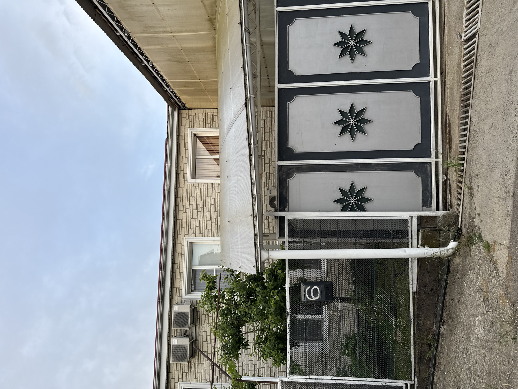 A vertical image shows the exterior of a building and a portion of the sidewalk, taken in Sochi, Russia. The building is a light beige brick structure with two visible windows on the side closest to the camera. Each window has a white frame. Two air conditioning units are mounted below the lower window. A white, semi-transparent awning is attached to the building. A metal gate with three decorative gray panels and black star-like designs spans the front of the building. Behind the metal gate, a mesh fence partially obscures a window. The number "6" is painted on a black rectangle attached to the fence. A small tree is visible between the fence and the building. A concrete sidewalk runs alongside the building, and a metal grate is embedded in the sidewalk. The sky above the building is pale blue with wispy clouds.