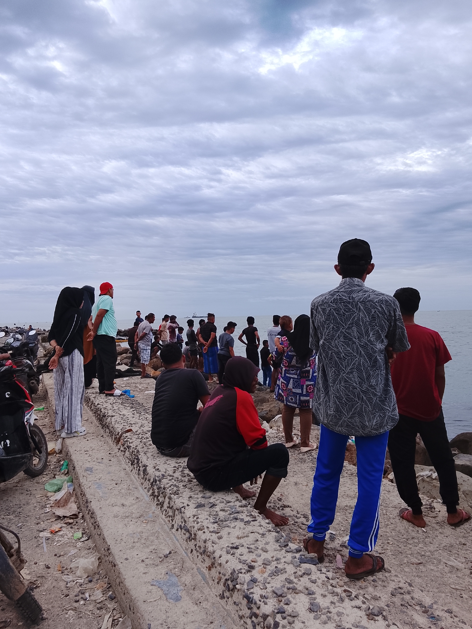 The image shows a group of people gathered on a concrete structure along the coast in Lhokseumawe, Indonesia. The sky is overcast. A mix of men and women are present. Some individuals are standing and looking out at the sea, while others are sitting. Clothing varies. Some people wear traditional Islamic attire. Several motorcycles are parked nearby. The area appears to be somewhat littered with debris. A cargo ship is visible on the horizon.