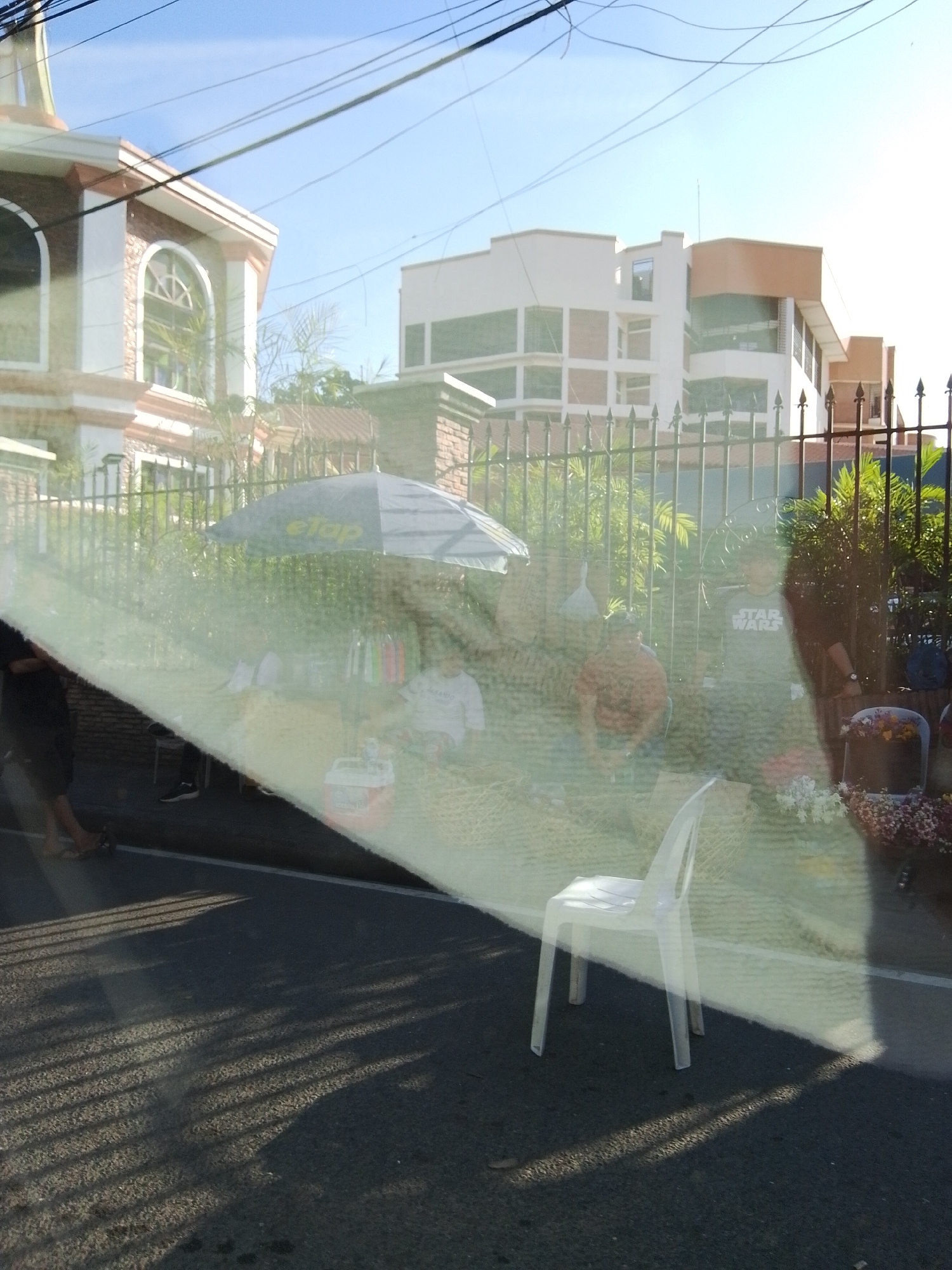 The image depicts an outdoor urban scene, likely taken through a reflective surface such as a window, as evidenced by a prominent diagonal glare spanning the mid-section and superimposed elements. The scene is consistent with an urban area in Davao City, Philippines, given the architectural styles and street vending presence.
In the background, on the left, a multi-story building with a light-colored facade (possibly white or cream) features reddish-brown accents and multiple arched windows, one of which displays a decorative criss-cross pattern. Above this building, a clear blue sky is visible, traversed by numerous dark power lines. On the right background, a more modern, multi-story building with a predominantly white exterior and some light orange-brown sections features an array of rectangular windows.
A black metal fence with pointed finials stretches across the mid-ground, separating the buildings from the foreground. Behind this fence, green foliage is interspersed. Several individuals are present in the mid-ground, somewhat obscured by the reflection. One person is seated under a dark umbrella bearing the "eTap" logo, indicative of a street vendor setup, with items hanging nearby and a red cooler on the ground. To the right of the vendor, another person is standing or seated, wearing a grey t-shirt with "STAR WARS" printed on it. Other indistinct figures are visible around this area.
The immediate foreground features a dark asphalt road with distinct, elongated shadows of vertical bars or a fence cast across it, suggesting a low sun angle. A single white plastic chair is positioned prominently in the lower-right foreground, also casting a shadow. On the far-right, partially obscured by the reflection, an arrangement of flowers or other decorative items is visible. On the far-left foreground, the lower legs and feet of a person are visible on the road surface.