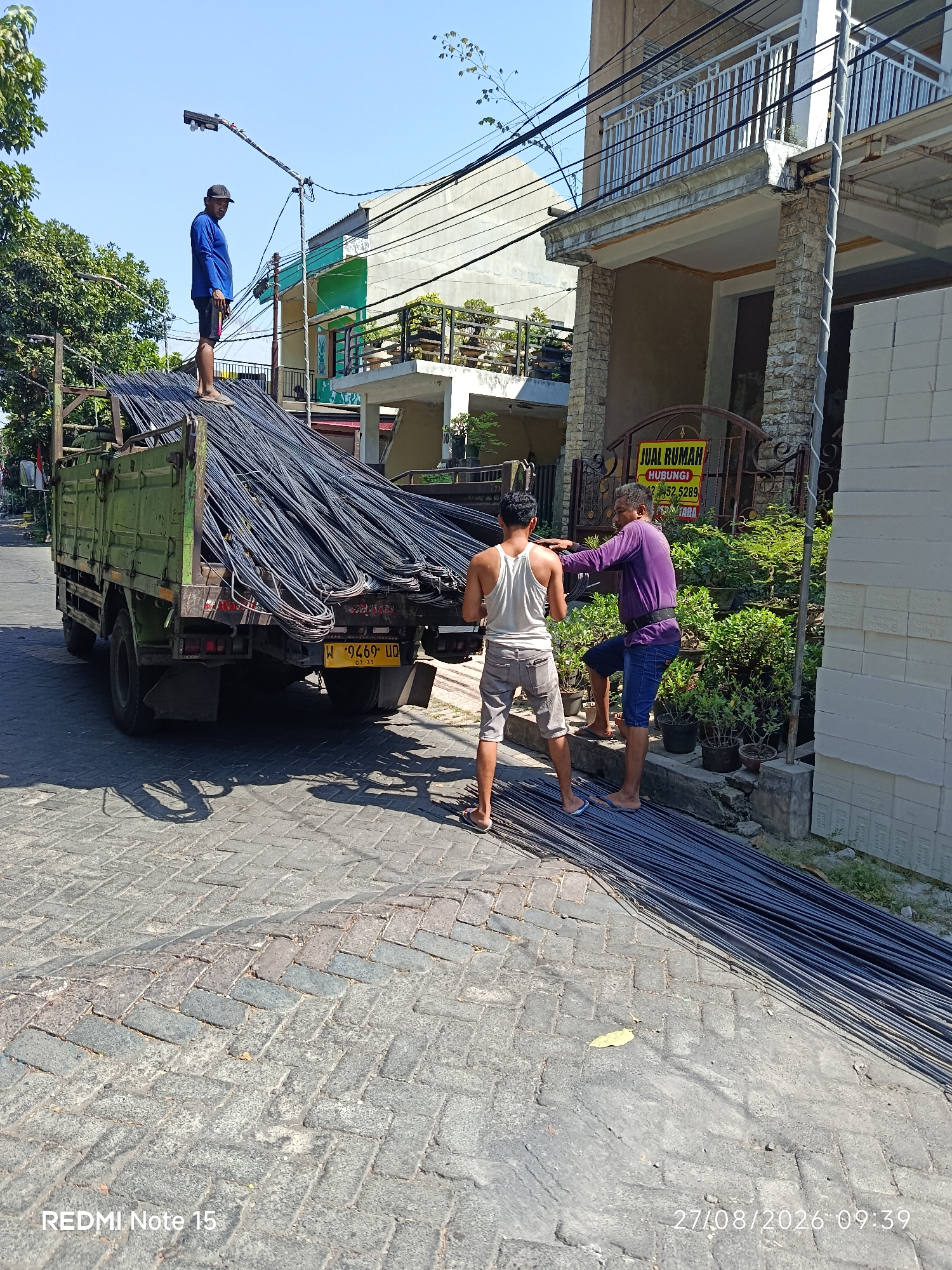 In Keboananom, Indonesia, a truck loaded with rebar is parked on a paved street. A man in a blue shirt and black cap stands on top of the rebar, looking towards the left. Two other men are on the street near the truck. One man, wearing a white tank top and grey shorts, is standing and holding a rebar. The second man, in a purple shirt and blue shorts, is leaning forward and appears to be talking to the man in the white tank top, with his hand on the man's shoulder. The rebar is also laid out on the ground in front of the truck, extending towards the right of the frame. Behind the men, a building with a balcony and a sign advertising "JUAL RUMAH" (House for Sale) is visible. Potted plants are placed along a low wall in front of the building. The sky is clear and blue, indicating a sunny day. The street is paved with interlocking bricks, and shadows from the truck and trees are cast on the ground.
