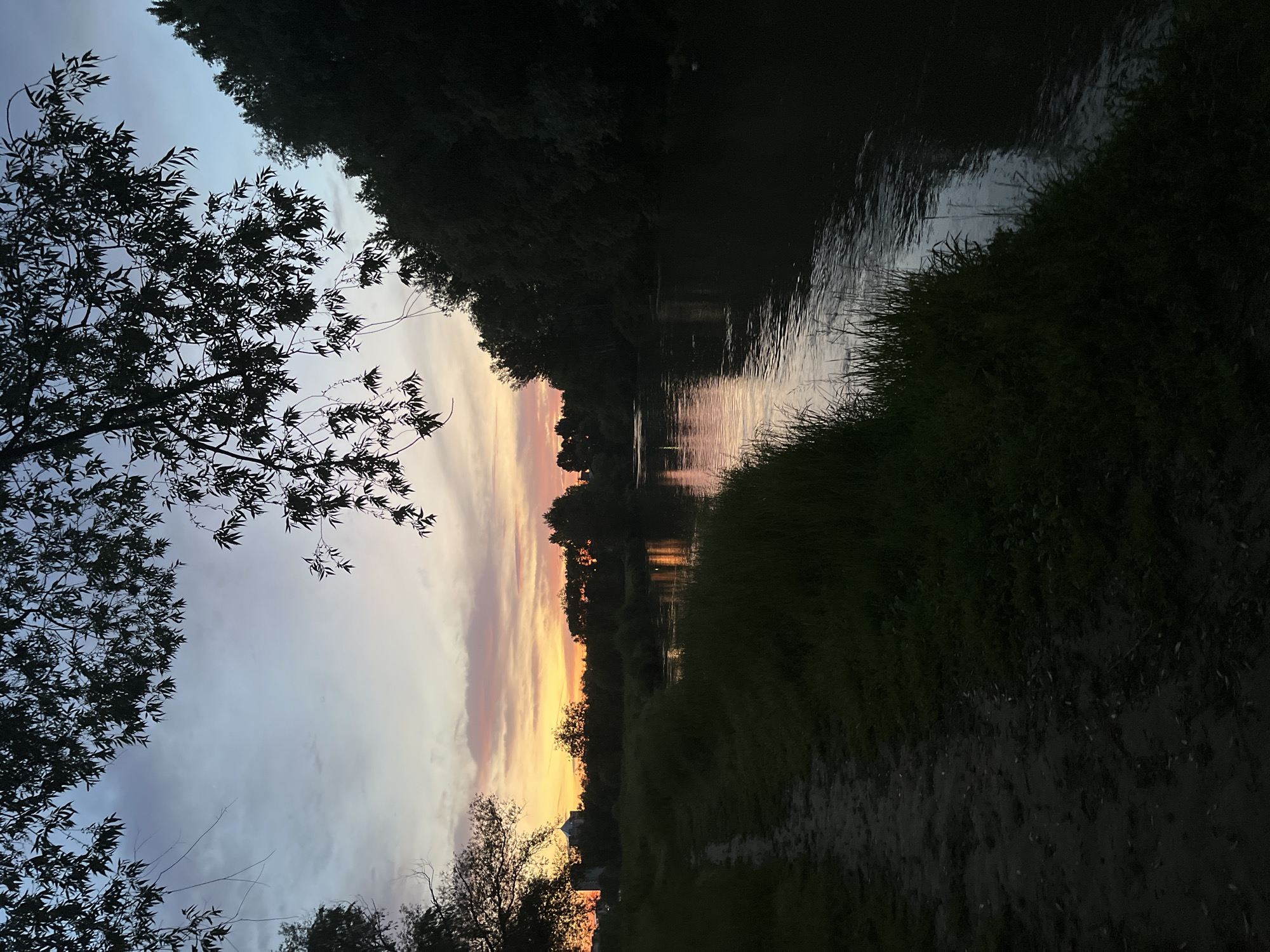 The image shows a body of water, likely a river, with a sunset reflected in its surface. On either side of the water are dark silhouettes of trees and foliage. A walking path is visible along the right side of the water. The sky contains shades of blue, pink, and orange. Based on the scenery, this location is potentially near the Maritsa River that flows through Plovdiv, Bulgaria. Without more information, the exact location within Plovdiv cannot be pinpointed. There are no people visible in the image.