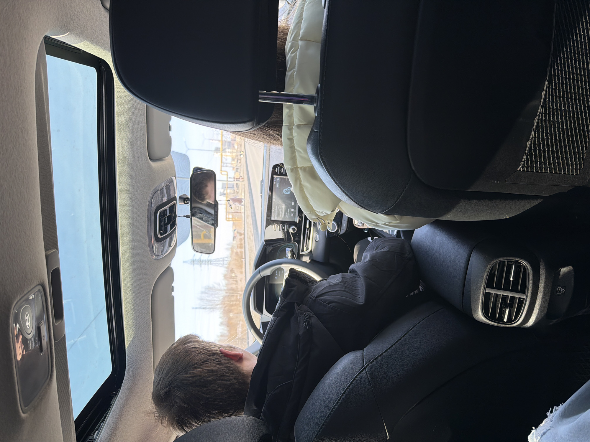 A person is seated in the driver's seat of a car. The interior of the car includes black leather seats, a dashboard with a screen, a steering wheel, and a sunroof. The rearview mirror reflects the passenger. The car is located in Berezniki, Russia. Outside the car window, power lines and industrial structures are visible. Another person is in the passenger seat, their jacket visible.