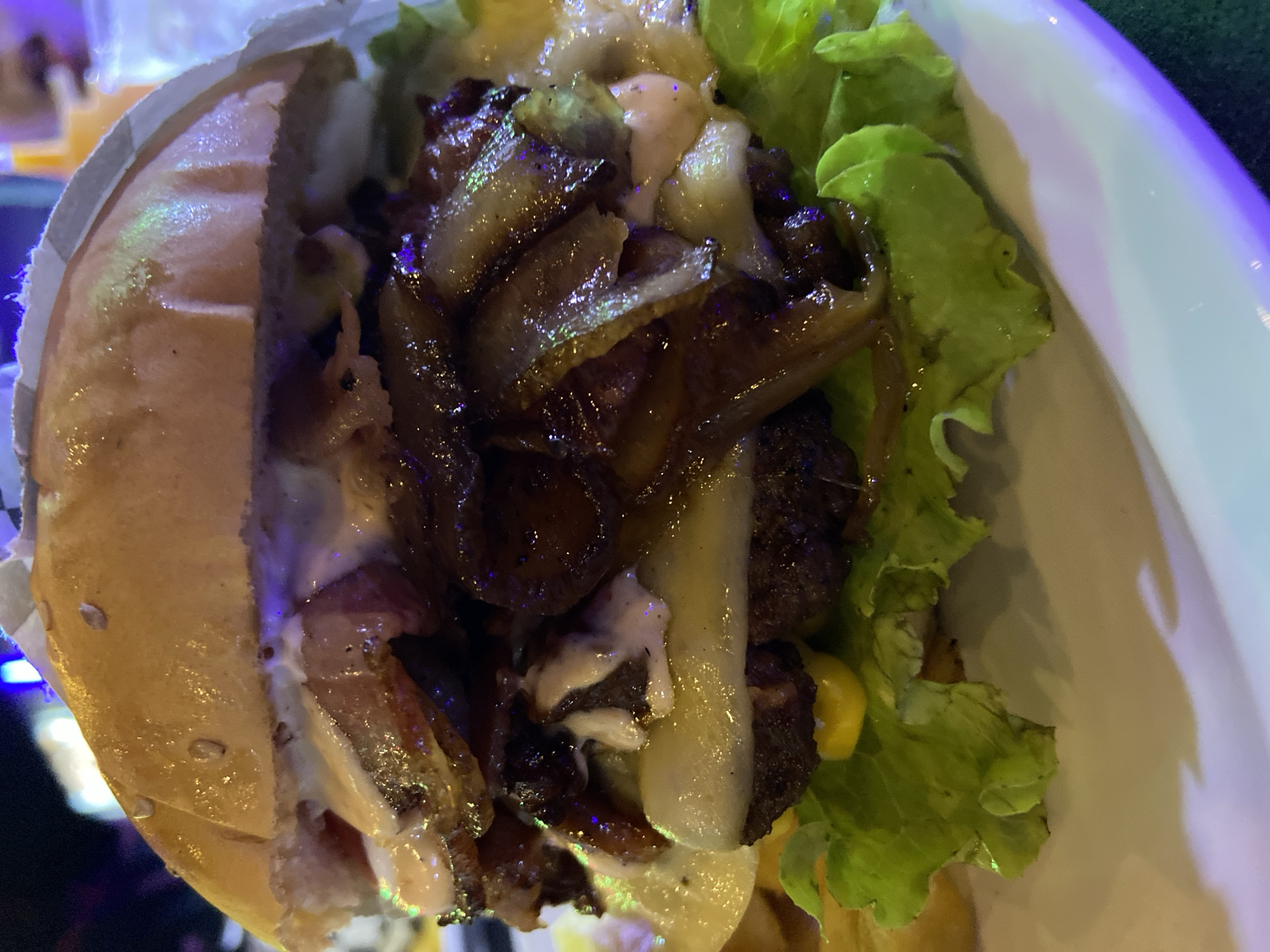 The image shows a close-up of a hamburger on a white plate. The hamburger has a sesame seed bun, lettuce, caramelized onions, sauce, and other fillings. The burger is partially wrapped in white paper. The lighting in the scene is a mix of cool and warm tones, suggesting artificial lighting. The location information is not discernible from the image.
