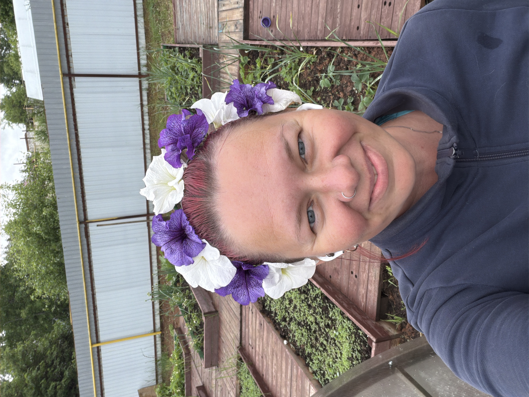 The image is a selfie taken outdoors. A person with fair skin, blue eyes, and a nose ring is in the foreground, wearing a flower crown of white and purple blossoms. The person is wearing a zipped-up blue garment. Behind the person, there are wooden garden beds with greenery growing in them. A white metal fence with a yellow horizontal bar runs along the left side of the frame, with green trees visible behind it. The scene appears to be in a garden or yard. While a precise location within the city of Chaikovsky, Russia cannot be determined solely from this image, the scene is consistent with a garden or outdoor area in that region.