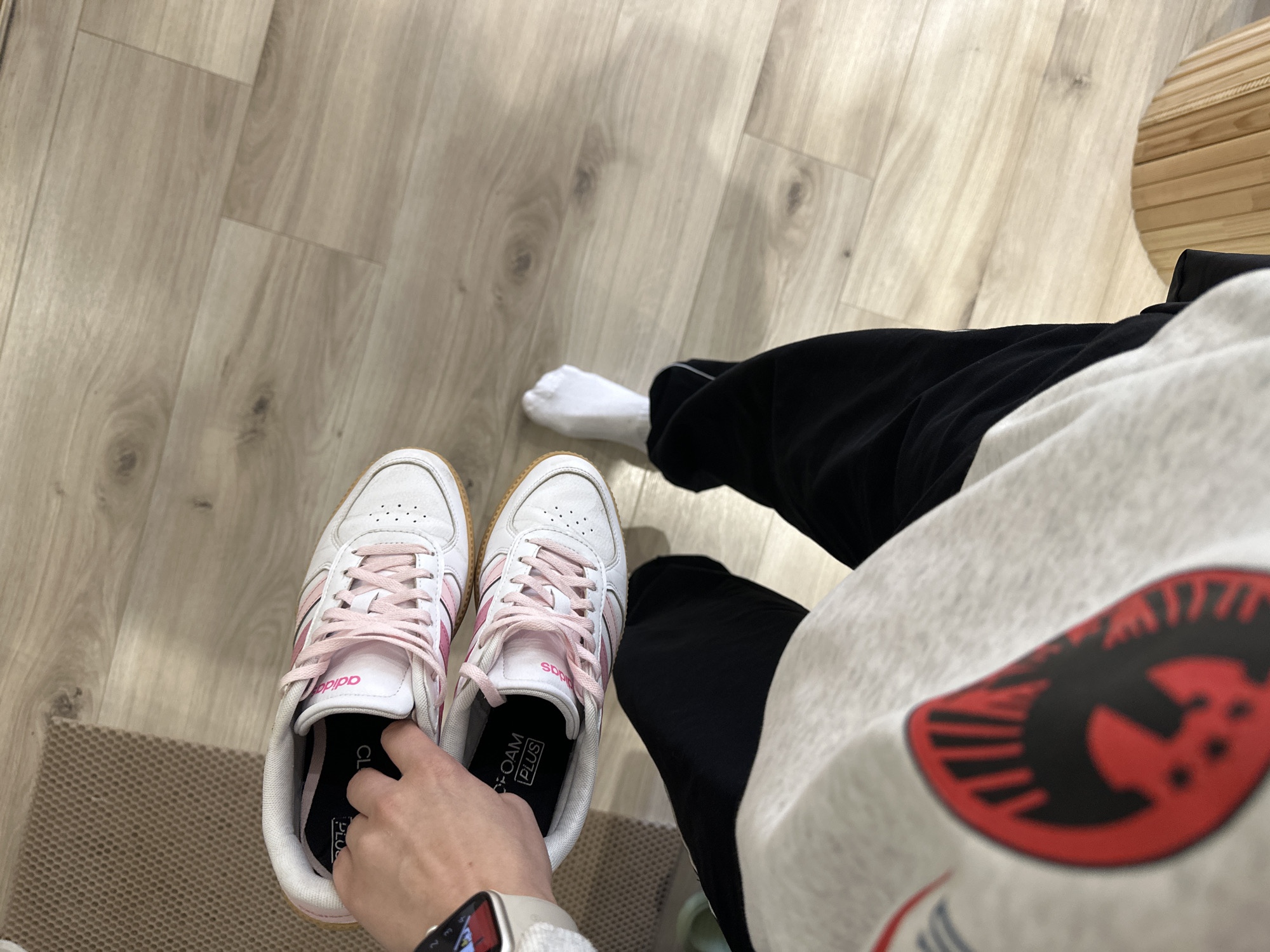 A first-person shot shows a person holding a white Adidas sneaker with pink stripes, against a light-colored wooden floor and wearing black sweatpants and a gray sweatshirt. A single white sock is visible on the right foot. The scene is possibly an indoor location and is in Perm, Russia given the user's request.