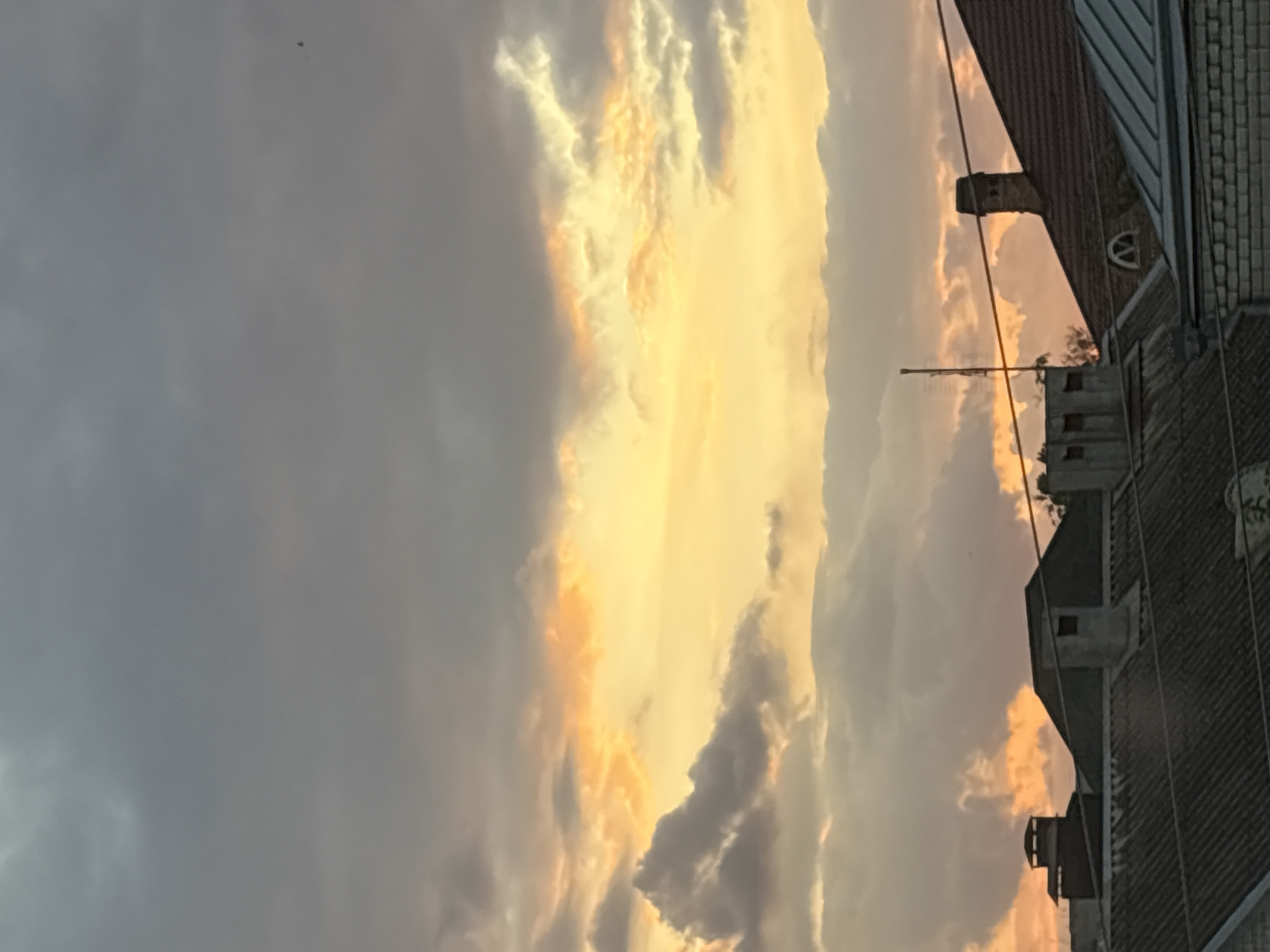 A low-angle, vertical shot captures a sunset over the rooftops of Georgiyevsk, Russia. The sky is filled with dramatic, billowing clouds painted in shades of gray, orange, and yellow, indicative of the golden hour. Several chimneys and rooftop structures are visible in silhouette against the vibrant sky, suggesting a residential or mixed-use area. The rooftops themselves appear to be made of corrugated metal and shingles. A thin antenna juts upwards from one of the buildings. A faint outline of what might be a bird is visible in the upper right portion of the sky. The overall impression is one of a tranquil evening scene in an urban environment.