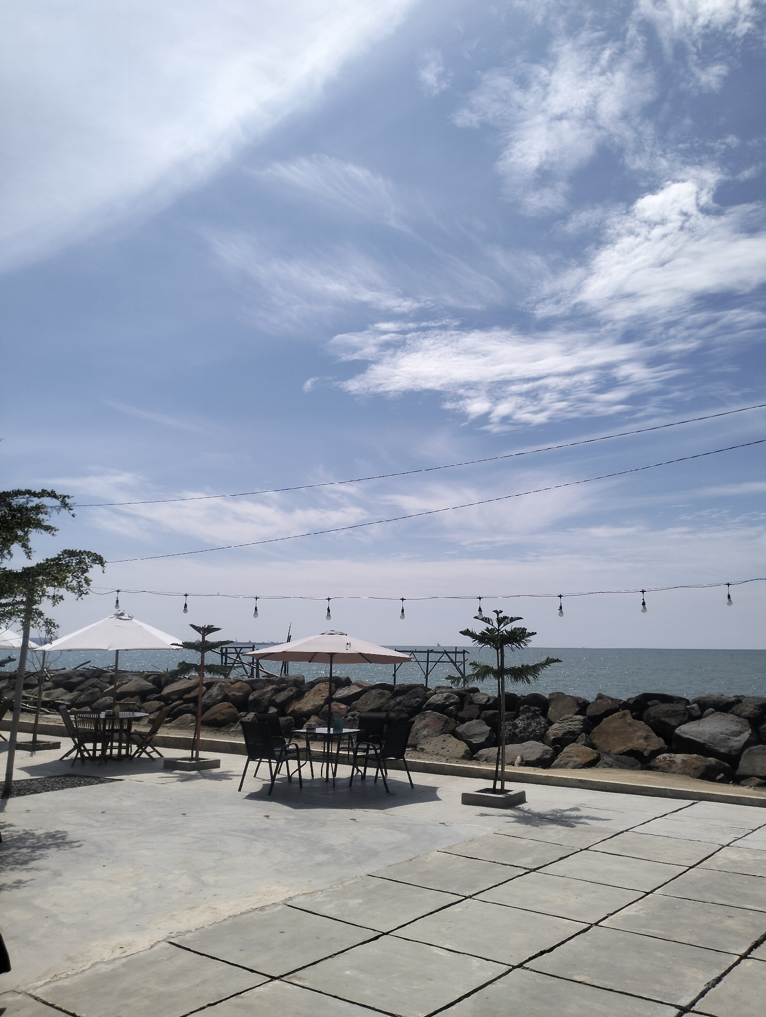 The image depicts an outdoor seating area by the sea in Lancok, Indonesia. The scene is characterized by a paved concrete patio with large square tiles, leading towards a rocky shoreline. Several tables and chairs are arranged on the patio, some of which are shaded by large white and beige umbrellas. String lights with exposed bulbs hang across the open space, suggesting it is a dining or relaxation spot.
Beyond the patio and rock barrier, the turquoise water of the sea stretches to the horizon under a bright blue sky dotted with wispy white clouds. A wooden pier or structure extends into the water in the distance. The overall atmosphere is serene and appears to be a calm, sunny day. There are no people visible in the frame.