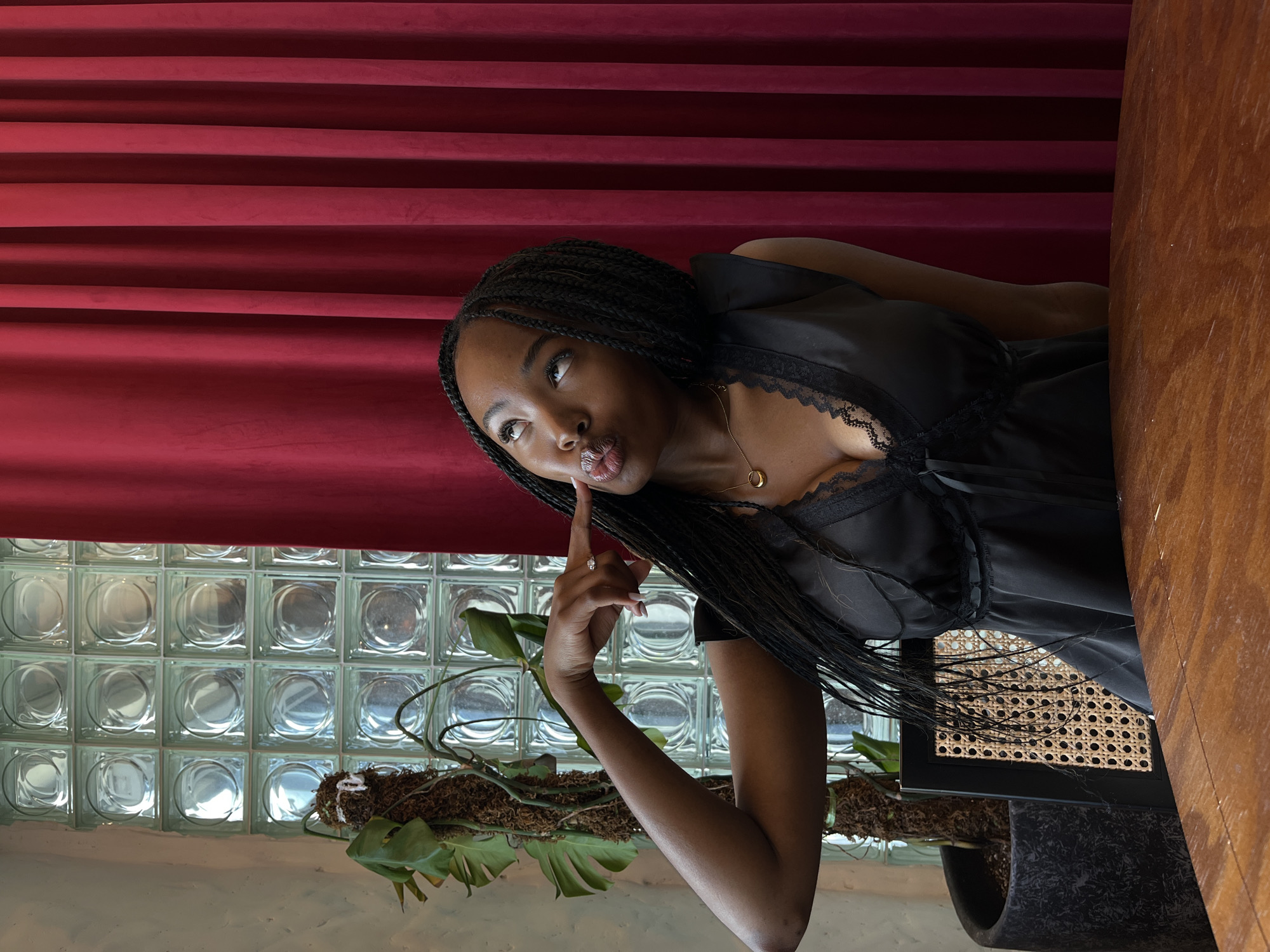 A young Black woman with long, braided hair is sitting at a wooden table. She is wearing a black slip dress with lace trim. Her right hand is raised to her face, with her index finger touching her cheek. She is looking upwards and to the right, with her lips pursed. The background consists of a wall of glass blocks and a red velvet curtain. A large potted plant is visible to the left of the glass blocks. A chair with a woven seat and back is partially visible behind the woman. The lighting is soft, creating a warm atmosphere.
