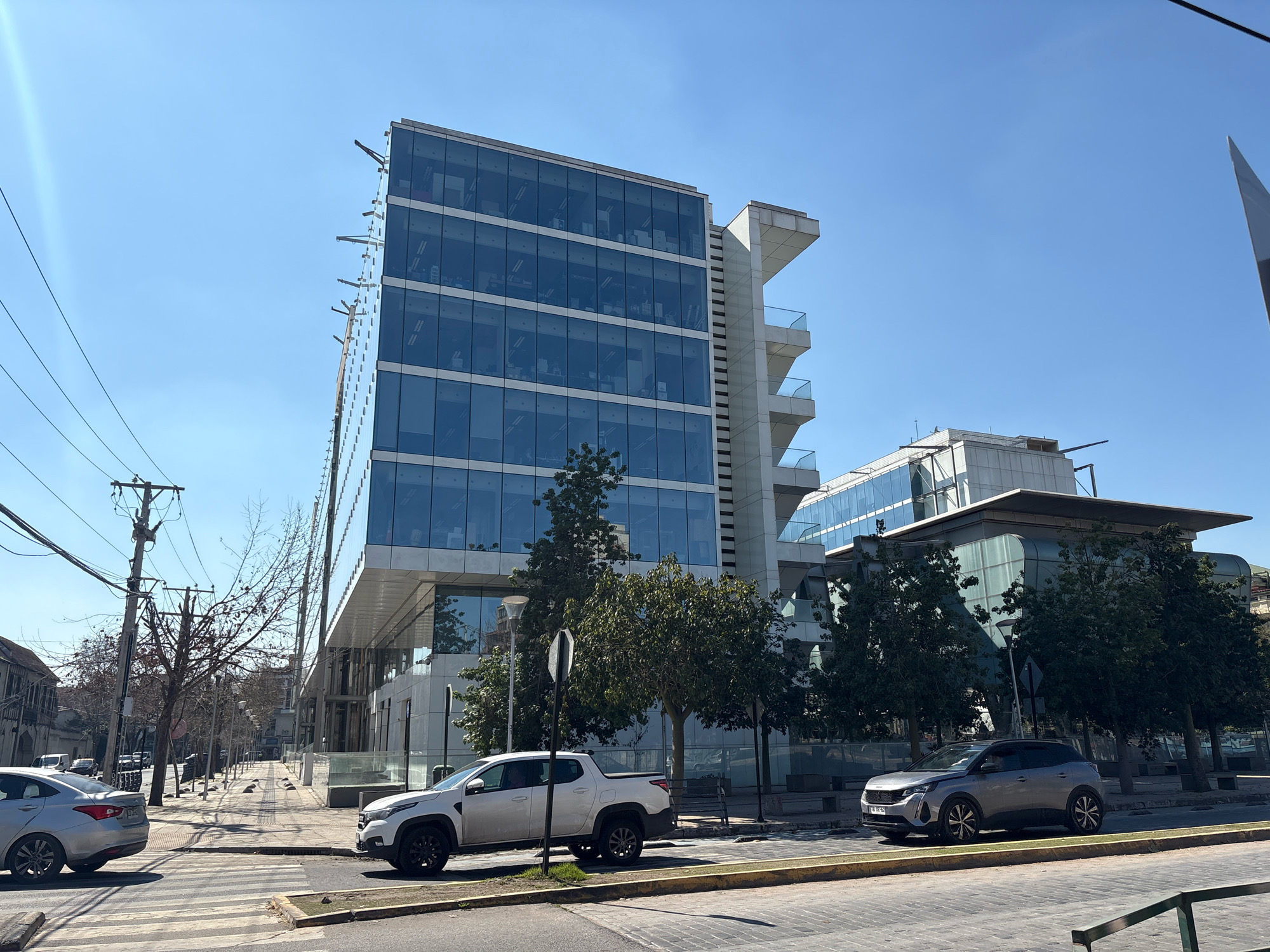 The image shows a street scene in Santiago, Chile, featuring modern architecture and urban infrastructure. A multi-story office building with a glass facade dominates the background. The building is sleek and contemporary, with multiple floors of reflective windows. To the right of the main building, another modern structure with a curved glass roof is visible.
In the foreground, two cars are parked on the street. A white pickup truck is on the left side of the road, and a silver SUV is on the right. Both vehicles appear to be stationary. A pedestrian crossing with yellow and white stripes is in the center of the image.
The street is paved with cobblestones in some areas and asphalt in others. Sidewalks are lined with trees, some of which are bare, suggesting a cooler season. Utility poles with numerous wires are present on the left side of the street, indicating the presence of electrical and communication infrastructure. The sky is clear and blue, suggesting a sunny day. The overall impression is of a well-developed urban environment in Santiago.
