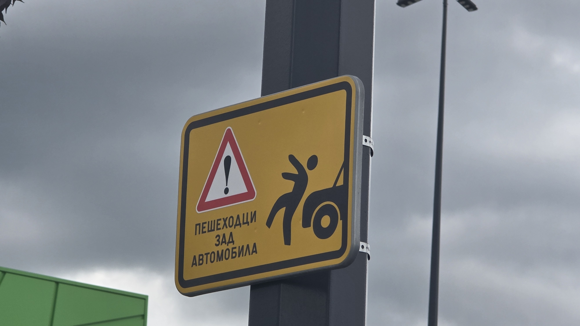 The image shows a yellow rectangular road sign with black text and graphics, attached to a dark gray metal pole. The sign depicts a pedestrian being hit by a car's rear. A red and white triangular warning symbol with an exclamation mark is also present. Below the graphics, text in Cyrillic script reads "ПЕШЕХОДЦИ ЗАД АВТОМОБИЛА". The pole is secured with two silver-colored metal bands. In the background, a light gray sky filled with clouds is visible, along with a tall lamppost and a partial view of a green building structure. The sign warns of pedestrians behind cars. The location is Pernik, Bulgaria, based on the Cyrillic text on the sign and the common use of this style of signage in Eastern Europe.