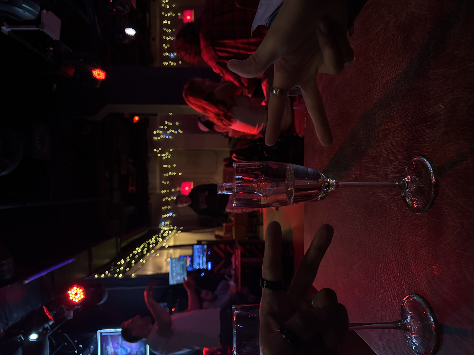 The image captures a scene at a bar, possibly in Moscow, Russia, characterized by a dark, red-lit ambiance, string lights, and a crowded environment. Two champagne glasses are centered on what appears to be a wooden table, with hands making peace signs above each glass, each hand wearing a ring. People are visible in the background; some are seated, and one appears to be dancing. In the background, a display of screens is visible. The lighting conditions are dim, with red and yellow accents.