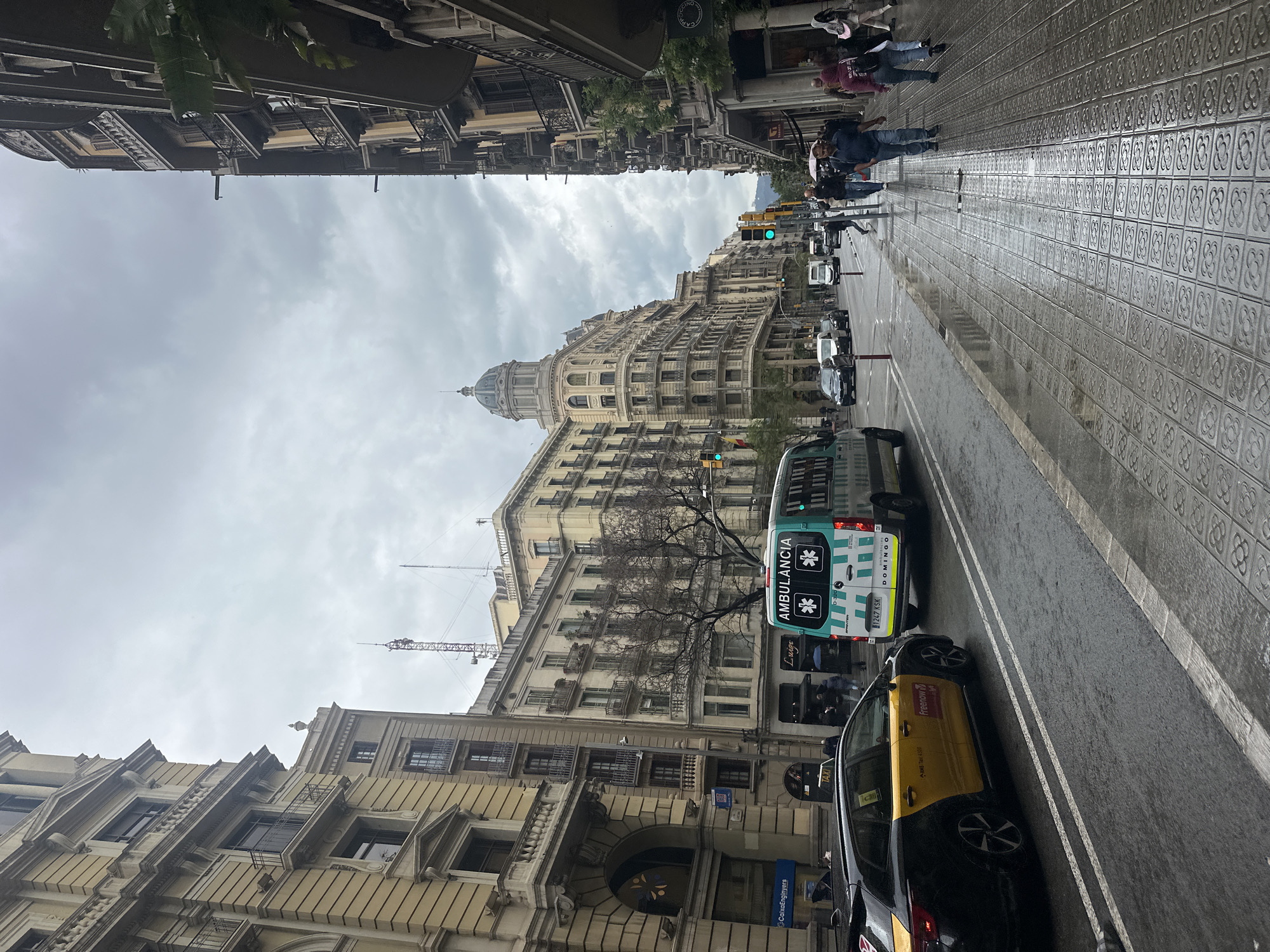 The image depicts a street in Barcelona, Spain, on a cloudy day, likely after a rain as the sidewalk and street are wet. Pedestrians are visible crossing the street. Cars, including a taxi and an ambulance, are parked along the road. The architecture is European with a mix of light-colored buildings. There is construction visible with a crane in the background. The sidewalk is paved with patterned tiles.