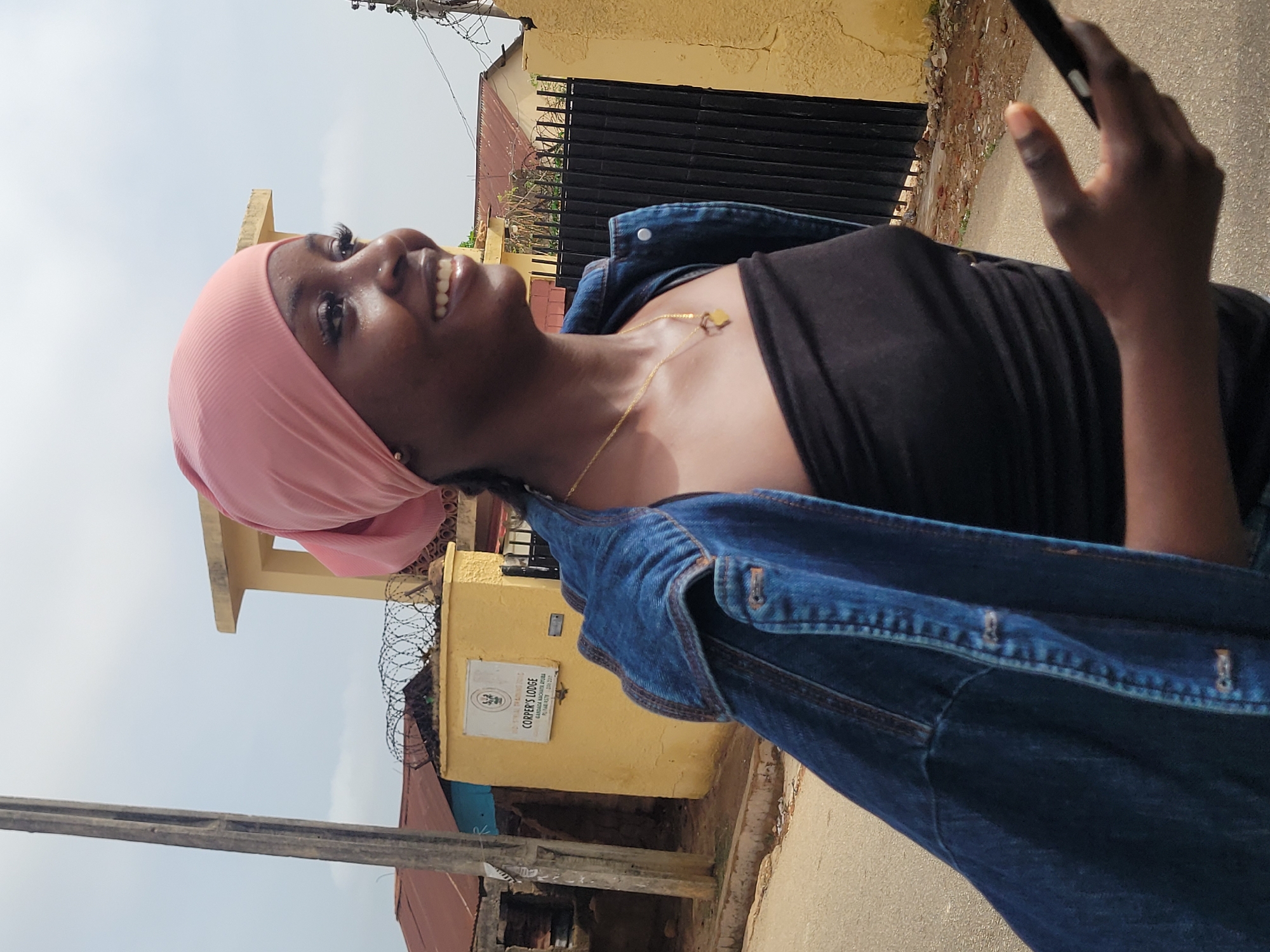 The photograph shows a young woman with dark skin smiling and looking upward. She is wearing a light pink head covering, a black top, and a blue denim jacket slung over her shoulders. She has a gold necklace around her neck and is holding a phone in her right hand.
The background includes a yellow building with a sign that reads "COPPER'S LODGE" with an address in Jos, Nigeria. Barbed wire is visible above the building. There's a black metal gate, and parts of other buildings with reddish roofs are also in view. The sky is overcast. The photograph appears to be taken outdoors on a street.