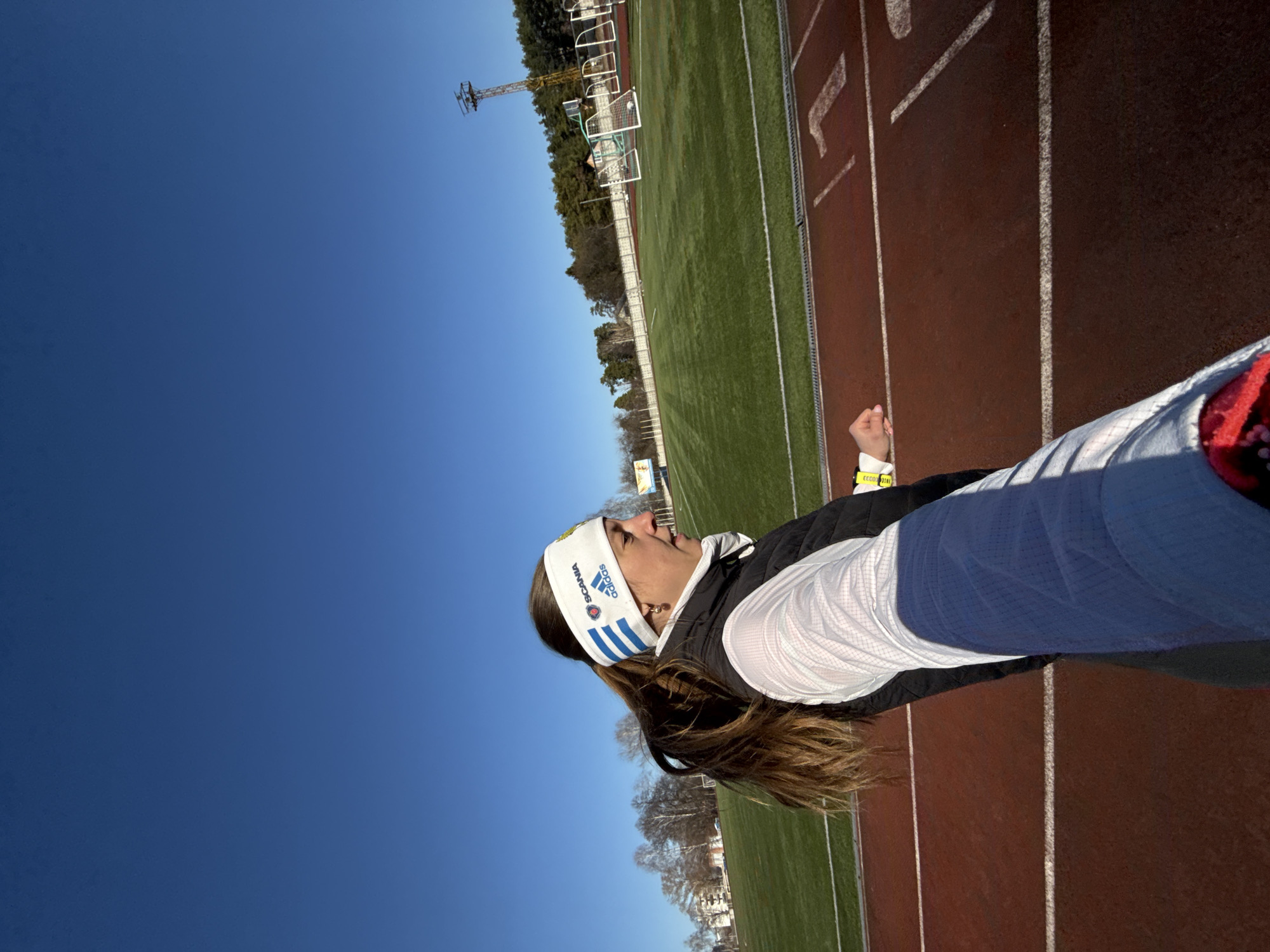The image captures a woman wearing a white Adidas headband and running attire on a red track, likely part of a sports stadium or training facility. The track is bordered by a green athletic field, and in the distance, a light tower and some goalposts are visible. Above, the sky is clear and blue. The photo appears to be taken in the city of Chaikovsky, Russia, based on the Adidas headband.