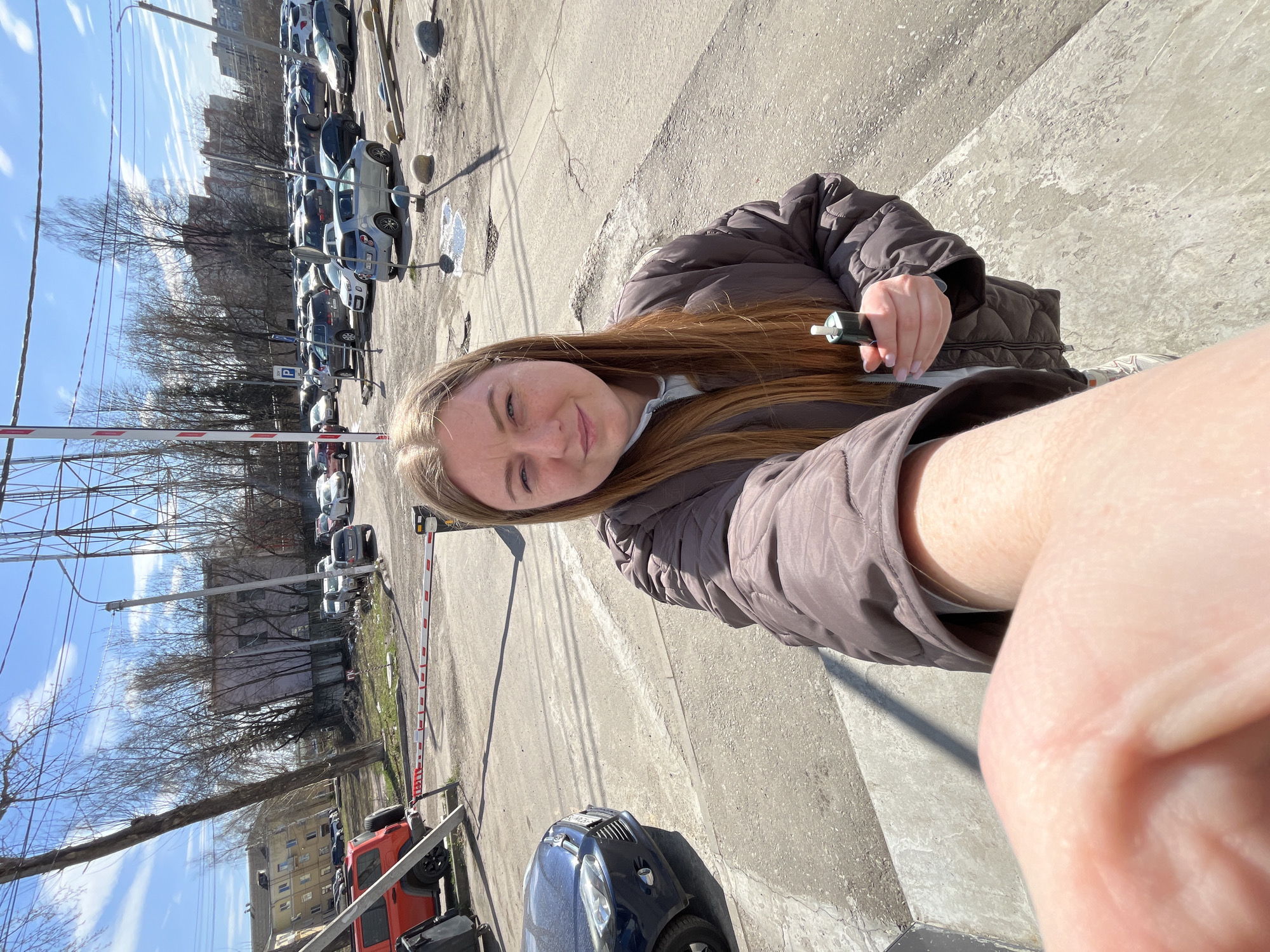 The image is a first-person perspective photograph taken in Klin, Russia, near a parking area and gate. The photo features a fair-skinned woman with light brown hair, wearing a brown puffer jacket, holding a small, dark-colored object in her hand, possibly a vape pen or similar item. The background includes a parking lot with numerous cars and a barrier gate. In the distance, buildings and bare trees are visible under a bright blue sky. The foreground shows part of the photographer's arm and hand. The setting suggests an urban environment, possibly near a residential or commercial area with parking facilities.