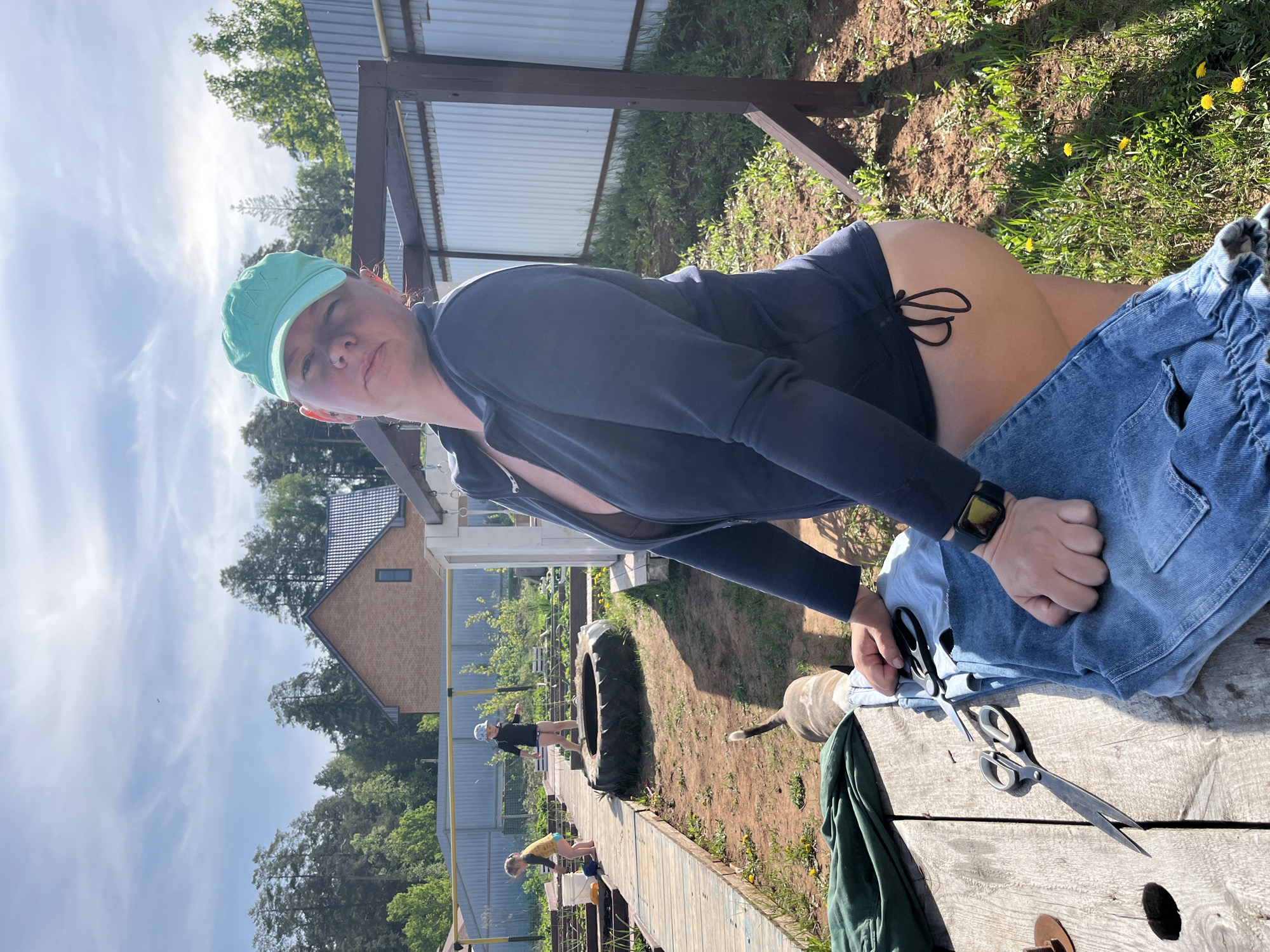 A woman wearing a blue cap and a dark blue top sits on a wooden table outdoors in Chaikovsky, Russia. She is working with a pair of scissors and blue jeans. On the table, there is also a green jacket or cloth. In the background, two children are playing near a tire and a small house can be seen. The area appears to be a yard with grass and trees.