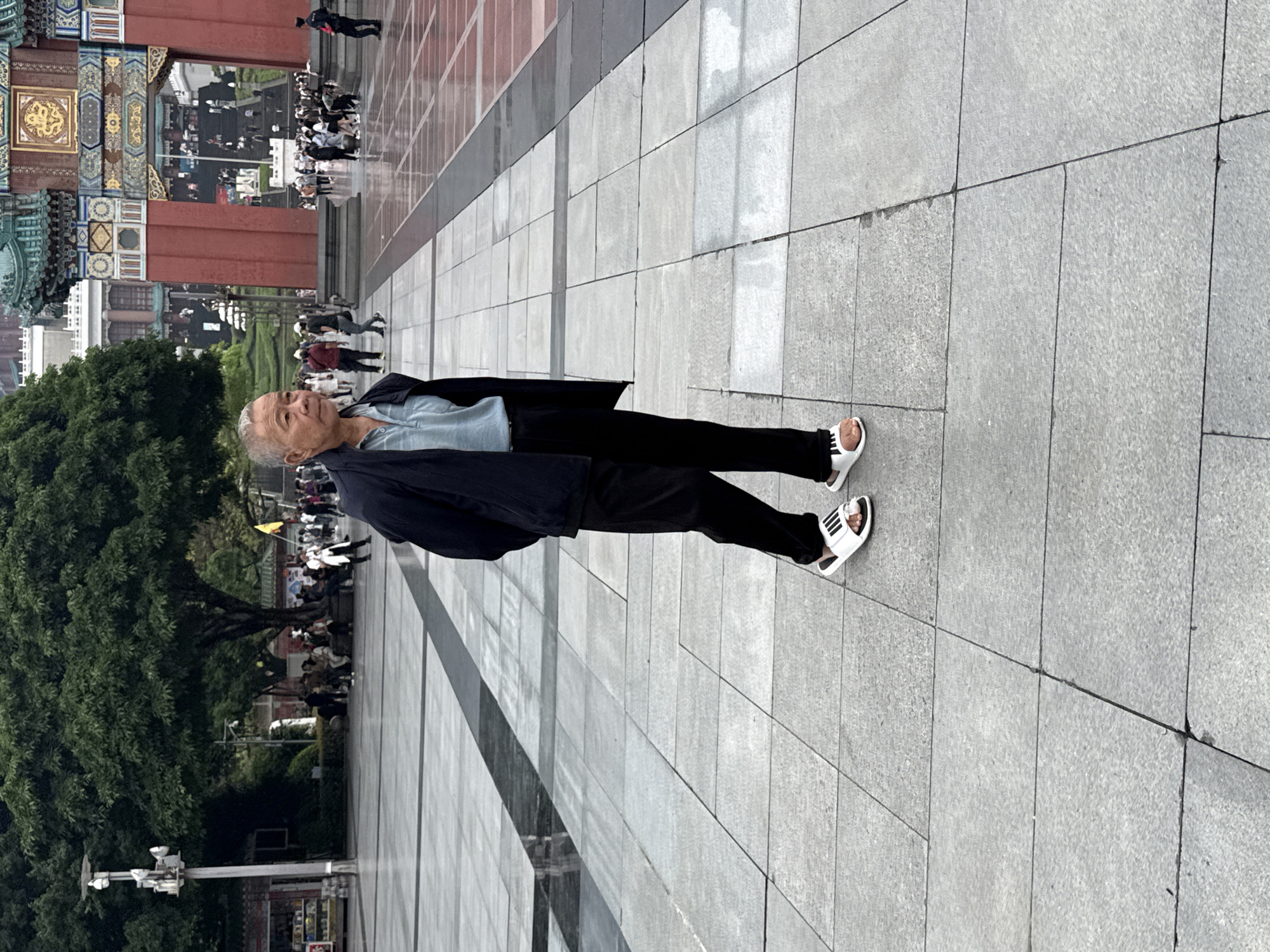 An elderly man stands on a paved walkway in Yuzhong District, China. He wears a dark jacket, light-colored shirt, dark pants, and white sandals with black stripes. In the background are trees, buildings, and other people. The paving stones of the walkway are light gray.