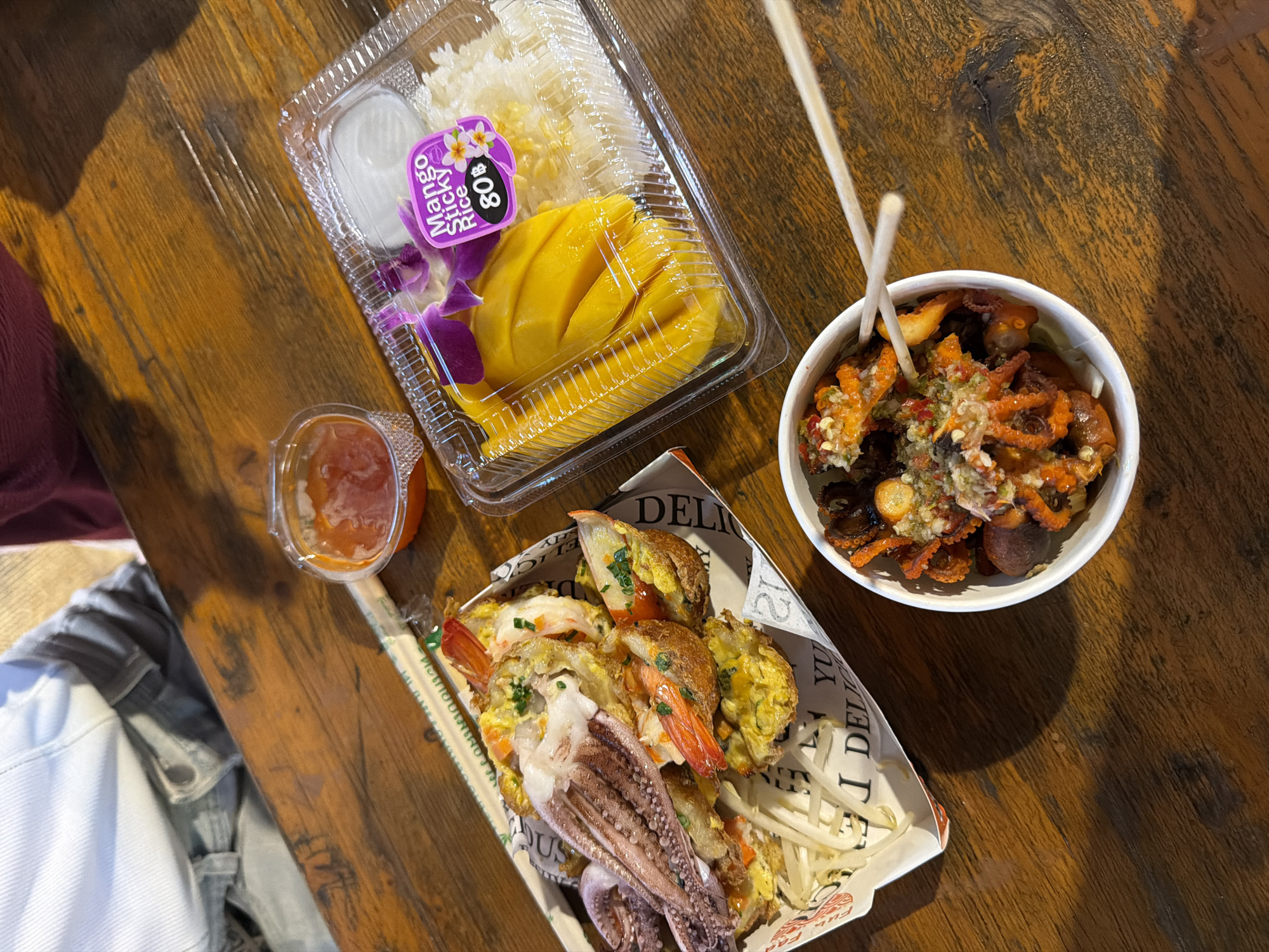 A close-up overhead shot shows a wooden table with several plates of Thai food in Bangkok, Thailand. In the foreground, a white bowl contains what appears to be crispy fried baby octopus tossed with a vibrant chili and herb dressing, with two chopsticks resting on top. To the left, a rectangular paper container holds a dish with battered and fried seafood, including a whole squid and several large shrimp, served over bean sprouts. A small plastic container of red chili sauce is visible next to it. Further to the right, a clear plastic container holds a portion of mango sticky rice, complete with sliced mango, sticky rice, and a small garnish of purple orchids. A sticker on the container reads "Mango Sticky Rice 80฿". The background shows a person wearing a light-colored denim jacket. The lighting suggests an indoor or shaded outdoor setting.