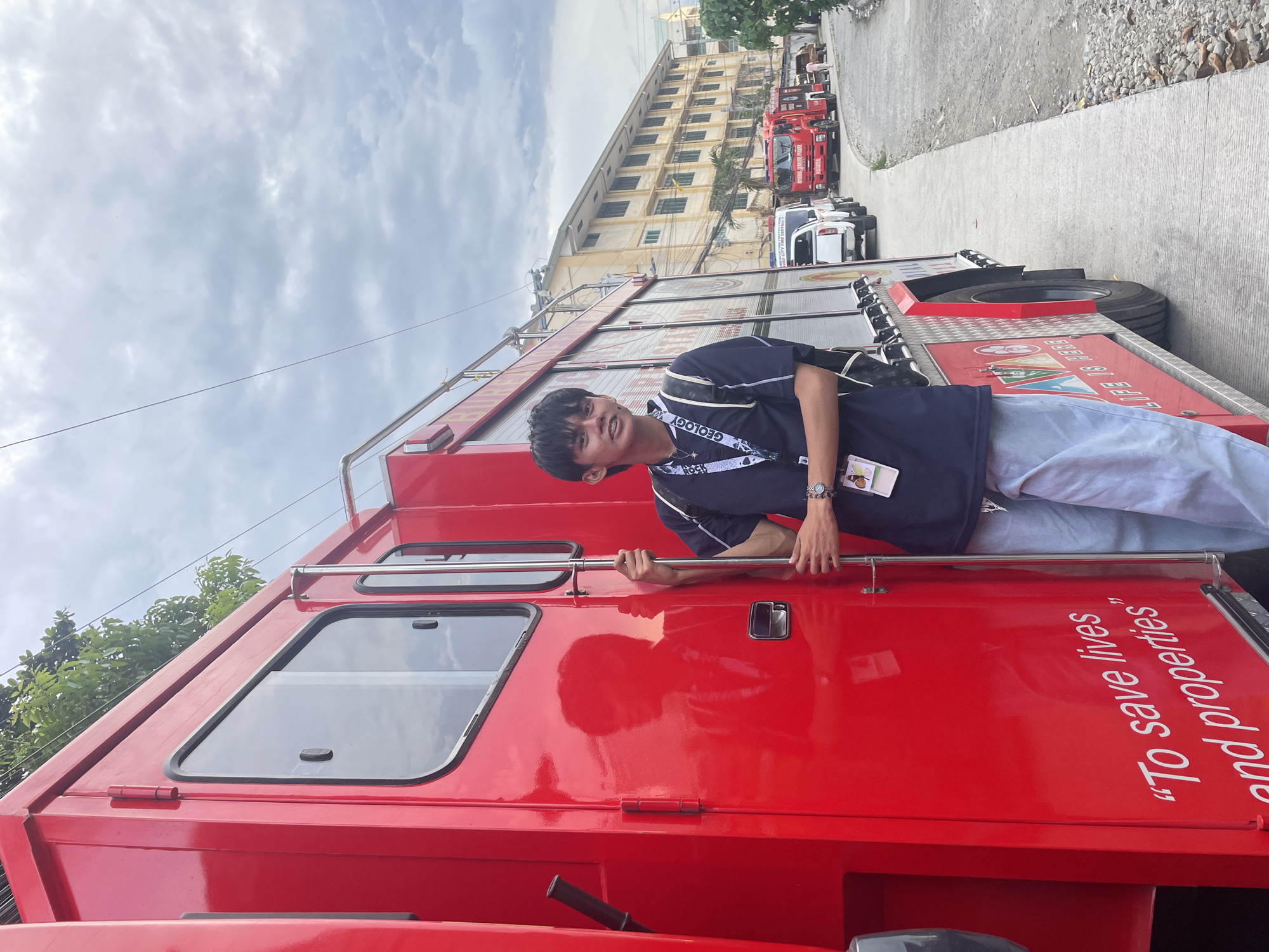 A young man stands next to a bright red fire truck, holding onto a metal pole. He is wearing a dark blue short-sleeved shirt with white trim, light blue jeans, and a backpack. He has dark hair and is looking towards the camera. The fire truck has "To save lives and properties" written on its side. In the background, several other vehicles, including another red fire truck and white pickup trucks, are parked along a street in front of a yellow building. The scene appears to be in Davao City, Philippines, as indicated by the text on the second fire truck in the background which reads "DAVAO CITY FIRE DISTRICT". The sky is cloudy.