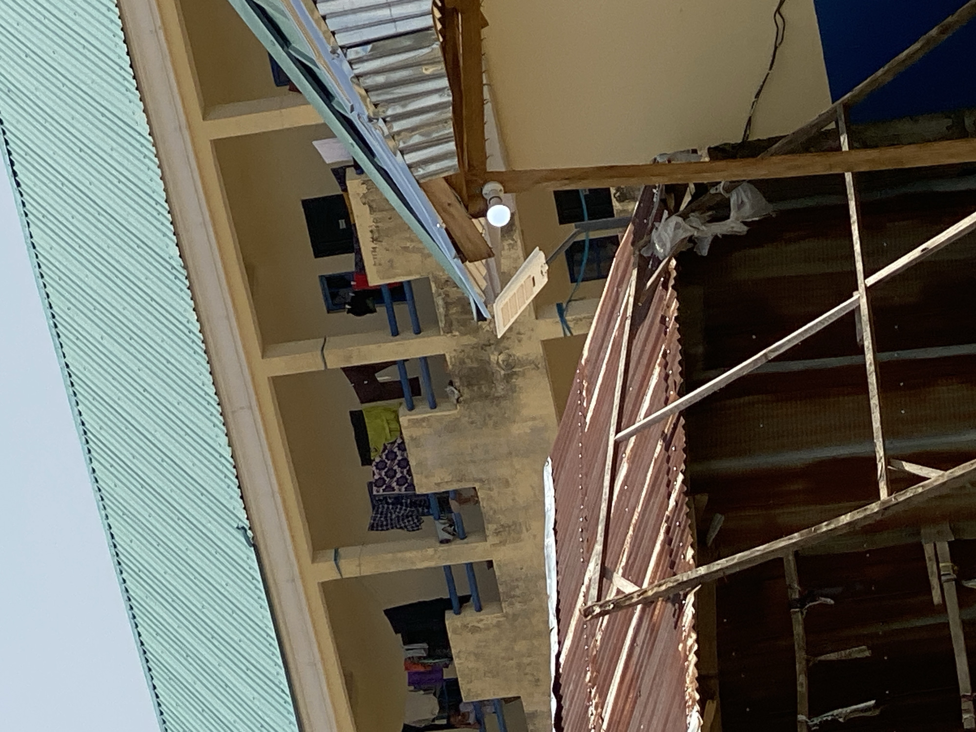 The image captures a scene with multiple levels of an outdoor building structure, possibly an unfinished construction or an older building in need of repair. On the left, a light blue corrugated metal roof runs diagonally across the frame, set against a clear sky. The main structure is a beige-colored building with open doorways or balconies on several floors. Blue railings are visible on these levels, separating them. Inside these open spaces, there are glimpses of what appear to be classrooms or rooms with desks and chairs. Some items, possibly clothing, are draped over the railings or placed on the desks.
The right side of the image features a rusted corrugated metal wall, suggesting an industrial or makeshift structure. Wooden beams and metal framing are exposed, with some pieces of white plastic sheeting caught within them. A single, exposed lightbulb hangs from a wire in the upper portion of the frame. The overall impression is one of an active, yet somewhat disorganized, outdoor environment. The scene is illuminated by natural light, implying it was taken during the daytime, with no clear indications of specific weather conditions. The setting appears to be in a warm climate, given the materials and open-air design. There is no visible text in the image.