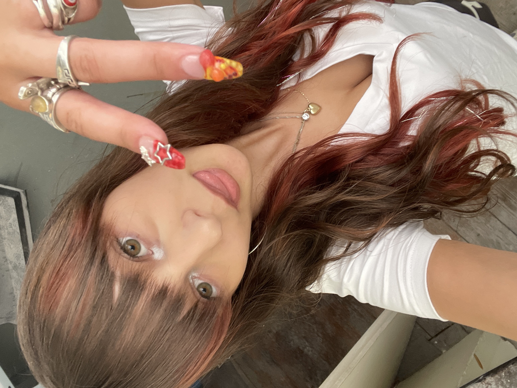 The image is a close-up, selfie-style shot of a young woman with brown hair with red highlights and bangs. She is wearing a white V-neck t-shirt and several rings on her fingers. Her fingernails are long and decorated with red and yellow designs. The background appears to be an indoor setting with wooden flooring and a blurred grey object. The image was taken in Raanana, Israel.