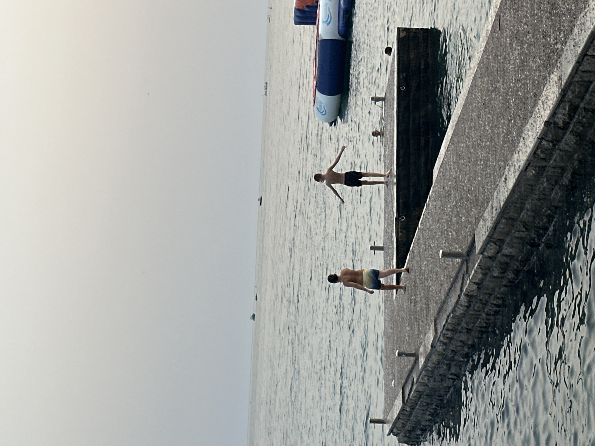 In Budva, Montenegro, on a clear day, two young boys are on a stone-lined pier extending into a calm, blue sea. One boy, wearing yellow and blue patterned swim trunks, stands facing away from the viewer, his back to the camera. The other boy, in dark swim trunks, stands further down the pier, arms outstretched as if preparing to jump or dive into the water. In the distance, several boats are visible on the horizon, and a large inflatable water structure is partially submerged in the sea to the right of the pier. The sky is a light gray, suggesting overcast conditions or early morning/late evening light.