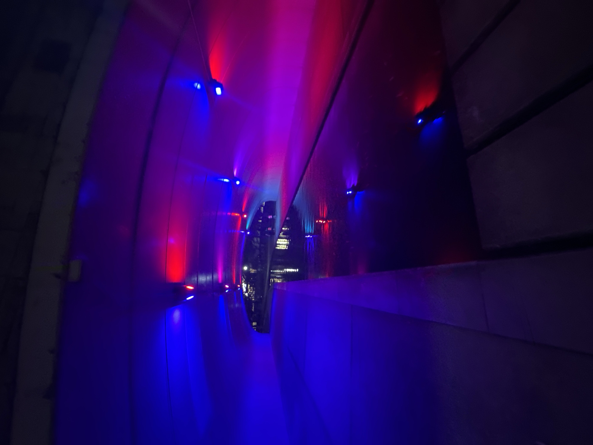 The image shows a tunnel under a bridge at night in Huntsville, Alabama. The tunnel is illuminated with a mix of blue and pink lights, creating a vibrant and colorful atmosphere. A canal or waterway runs alongside the walkway, with reflections of the lights visible on the water's surface. The walkway appears to be made of concrete. In the distance, the lights of the city are visible, along with silhouettes of people walking. The scene suggests a public space that is lit up at night for aesthetic purposes or safety.