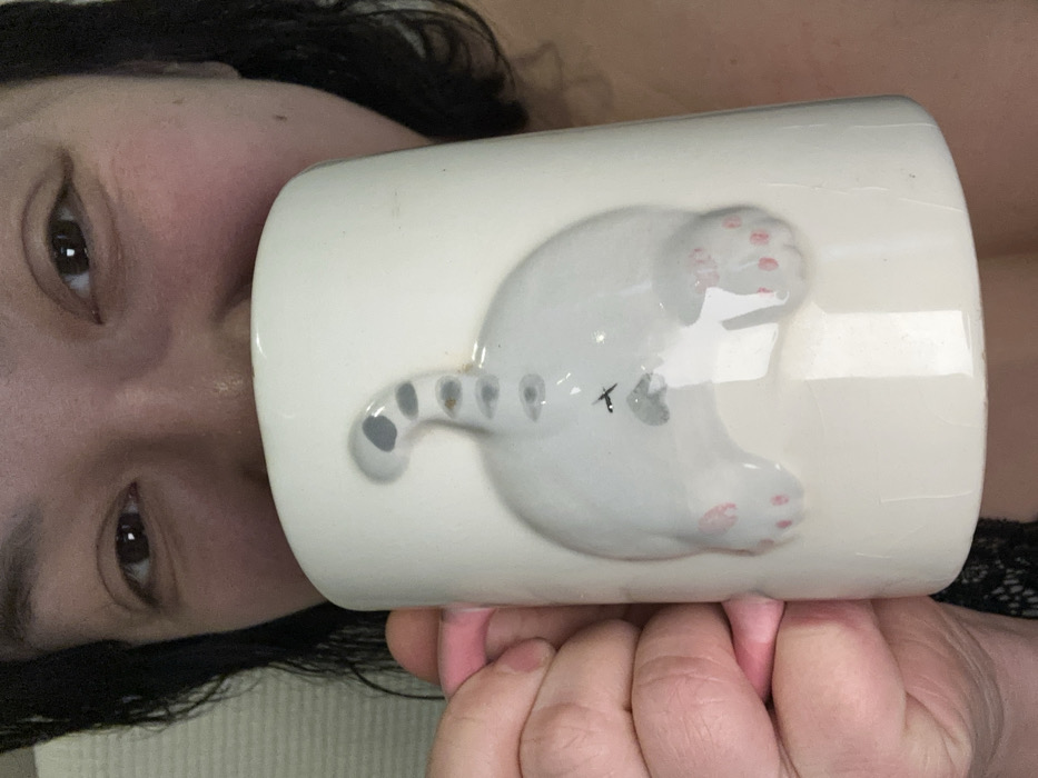A woman's face is visible alongside a white mug. The mug features a sculpted, grey cat design, showing its rear end with paws and a spotted tail. The woman's brown eyes are prominent, and her dark hair frames her face. She appears to be holding the mug with both hands, fingers slightly visible. A hint of dark lace clothing is present near her shoulder.