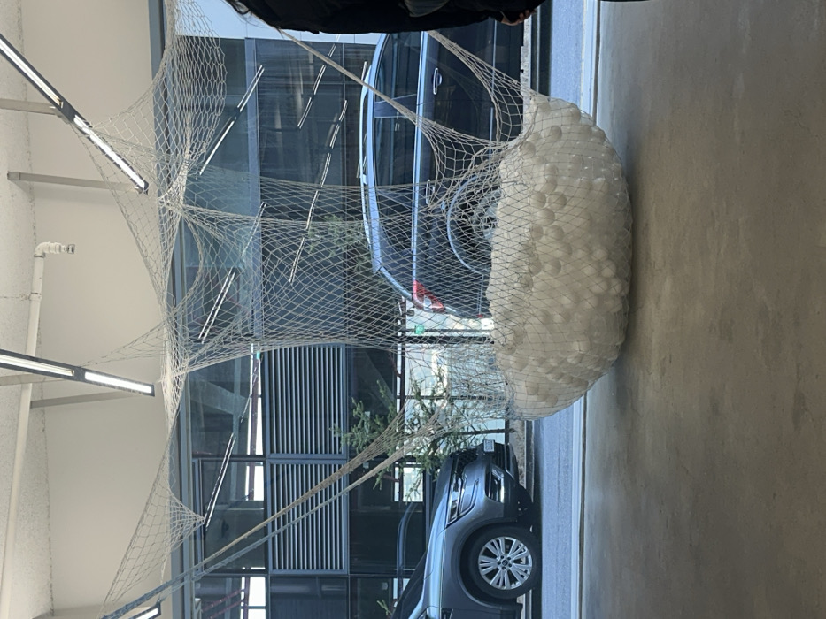 The image presents an interior view looking through a large glass window onto an urban street in Seoul, South Korea. A contemporary art installation dominates the indoor foreground, consisting of an extensive white net structure suspended from the ceiling. This netting forms a large, irregular pouch-like receptacle filled with numerous small, uniform white spherical objects, resting approximately one-third of the way up from the floor. The indoor floor is a light grey concrete surface. The ceiling displays exposed white pipes and multiple linear fluorescent light fixtures.
Through the window, a street with vehicles is visible. On the left, a silver-grey Renault XM3 or Arkana SUV is parked or stationary, facing right. To its right, a dark grey or black SUV is partially visible, facing left. The background features modern building facades with horizontal louvers and glass panels, alongside sparse leafless trees or bushes. Reflections of the indoor ceiling and lighting are apparent on the windowpane. On the far right edge of the frame, a partial figure, dressed in dark clothing, is visible indoors. No discernible actions or interactions are occurring.
