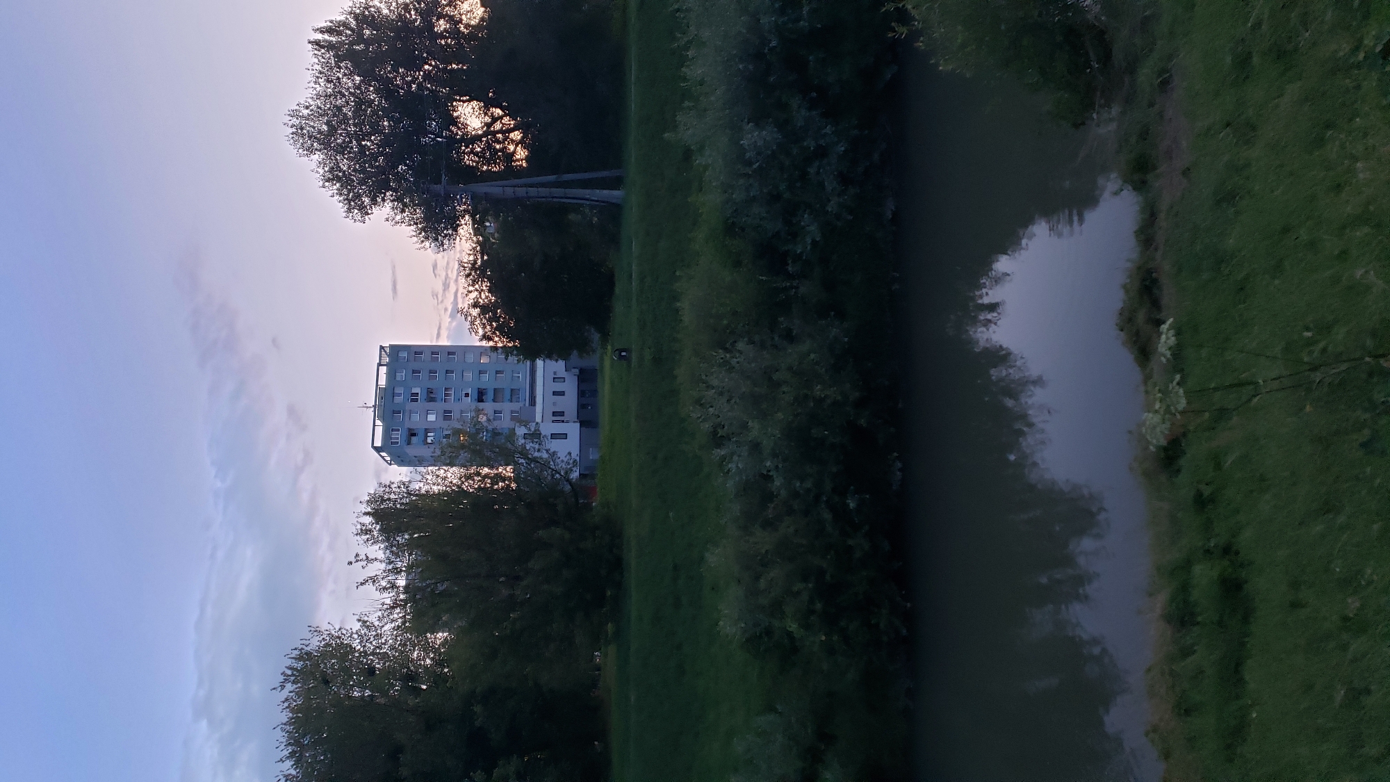 The image depicts a twilight scene in Prešov, Slovakia, focusing on a residential apartment building situated on a grassy embankment overlooking a calm body of water. The building, a multi-story structure with a light blue facade, features numerous windows and a flat roof with visible antennas. The surrounding area is characterized by lush green grass and dense foliage from various trees, some of which are silhouetted against the subtly colored sky, which transitions from a pale blue at the top to soft pinks and oranges near the horizon. The water in the foreground reflects the trees and the sky, creating a tranquil and somewhat melancholic atmosphere. A utility pole with wires stands to the right of the building, partially obscured by trees. There are no people or animals visible in the frame.