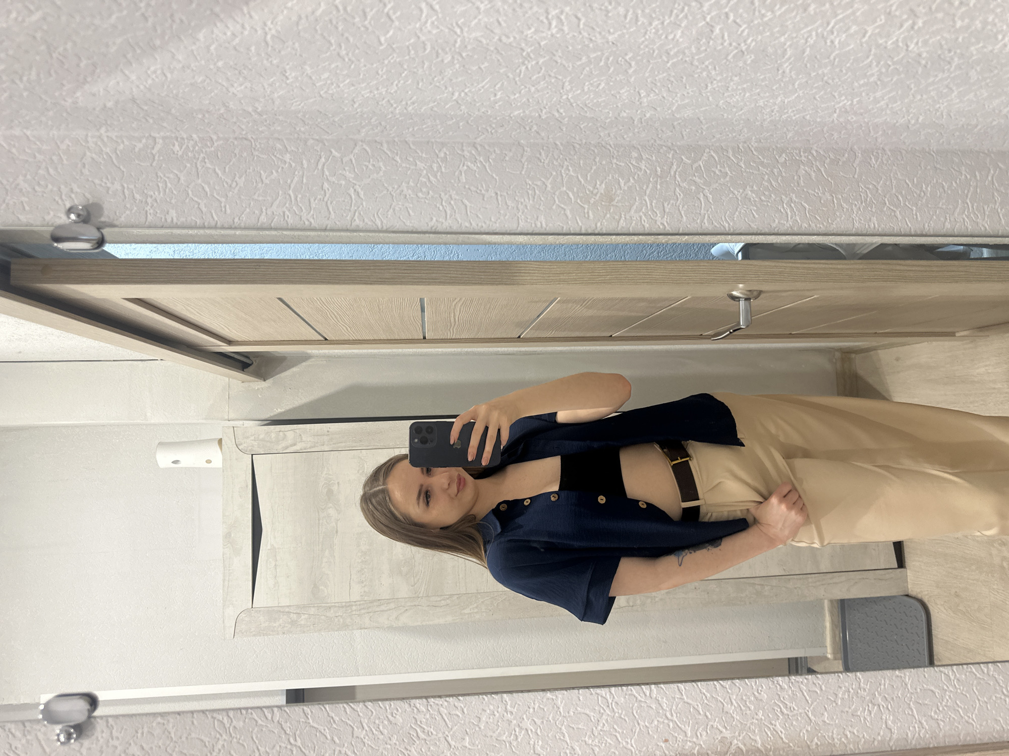 A woman is taking a selfie in front of a mirror. She is wearing a black top, a navy blue button-down shirt, and tan pants with a brown belt. The background includes a white textured wall, a cabinet with a tissue dispenser, and a light wood-colored doorframe. The location is in an unknown city.
