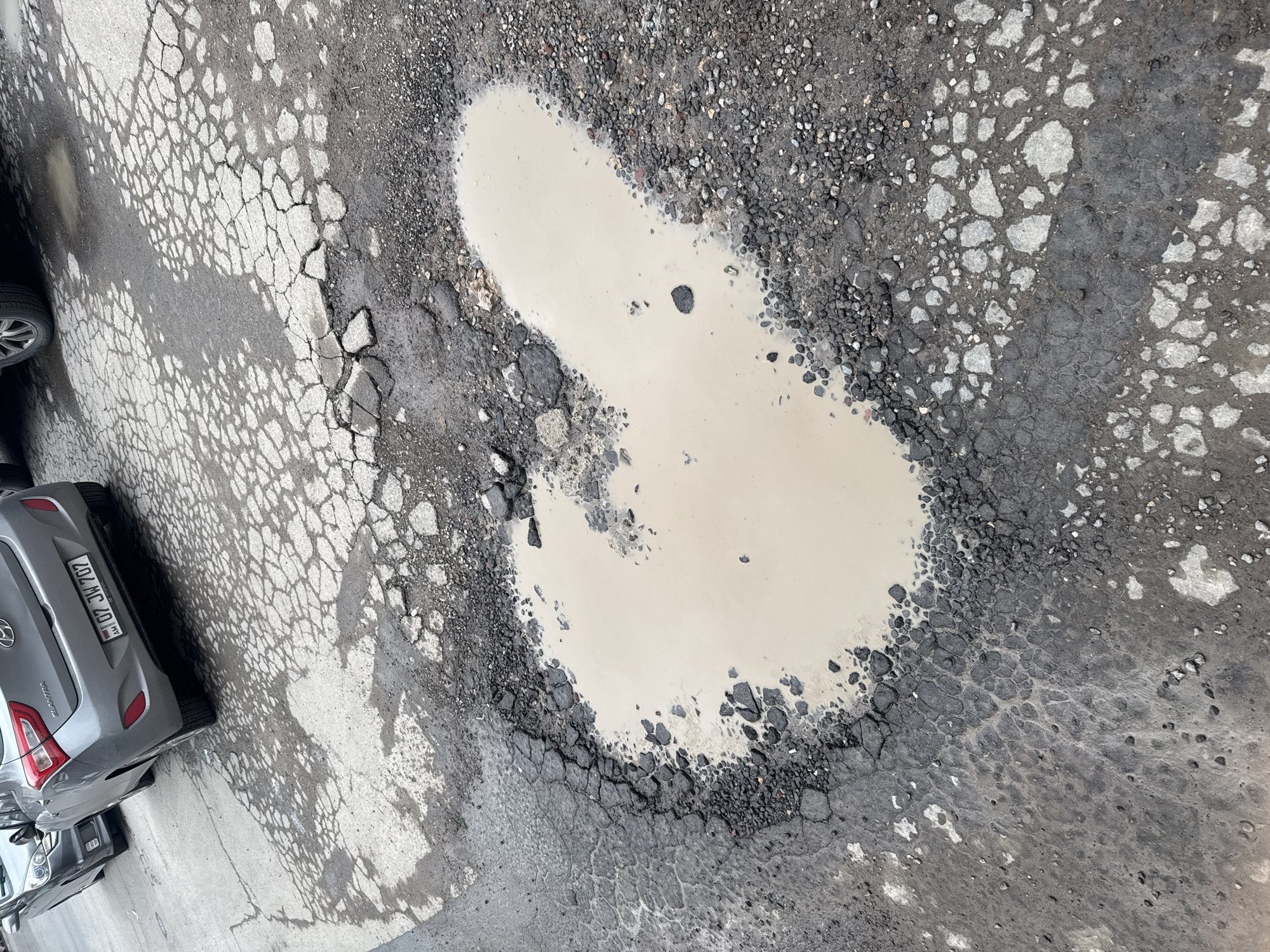 The image shows a water-filled pothole in a cracked and damaged asphalt road surface. The pothole is surrounded by loose gravel and broken asphalt chunks. Adjacent to the damaged road is a parked silver Hyundai Elantra, with a license plate that appears to read "707 MC 40 | AM." The location is suspected to be Yerevan, Armenia due to the format of the vehicle registration plate on the car.