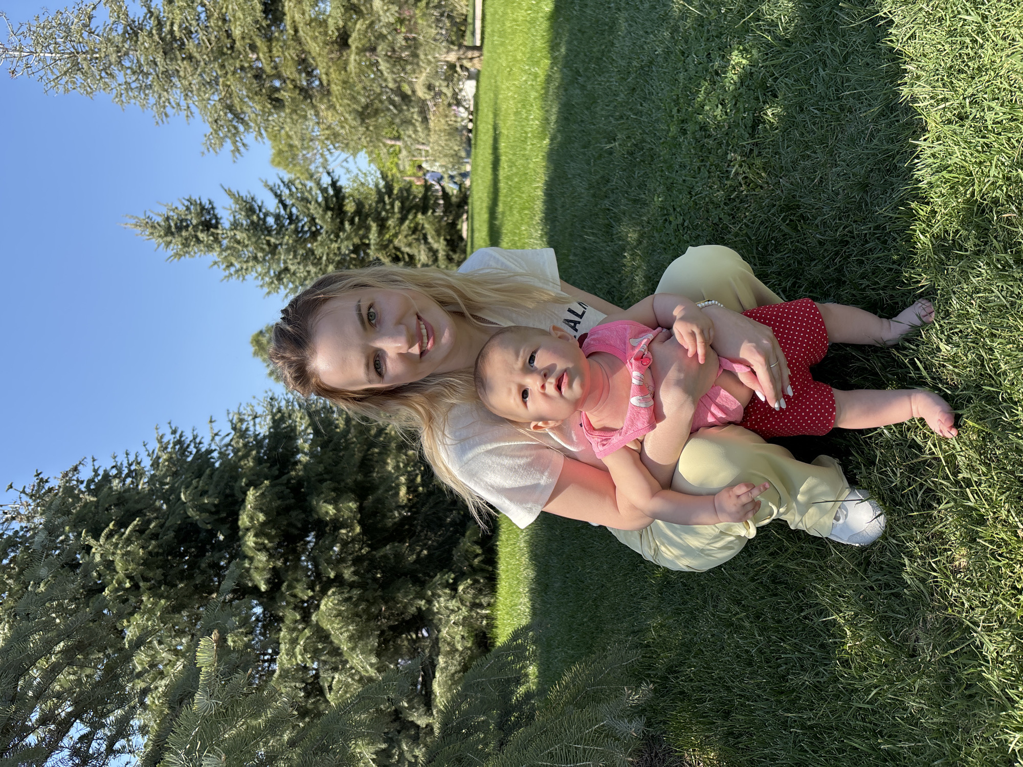 A woman sits cross-legged on a lawn, holding a baby. The woman has blonde hair and is wearing a white t-shirt and yellow pants, and white shoes. The baby is wearing a pink top and red and white polka dot bottoms. The setting appears to be outdoors in a park or green space. Large evergreen trees are visible in the background. The city of Krasnodar, Russia, is unspecified.