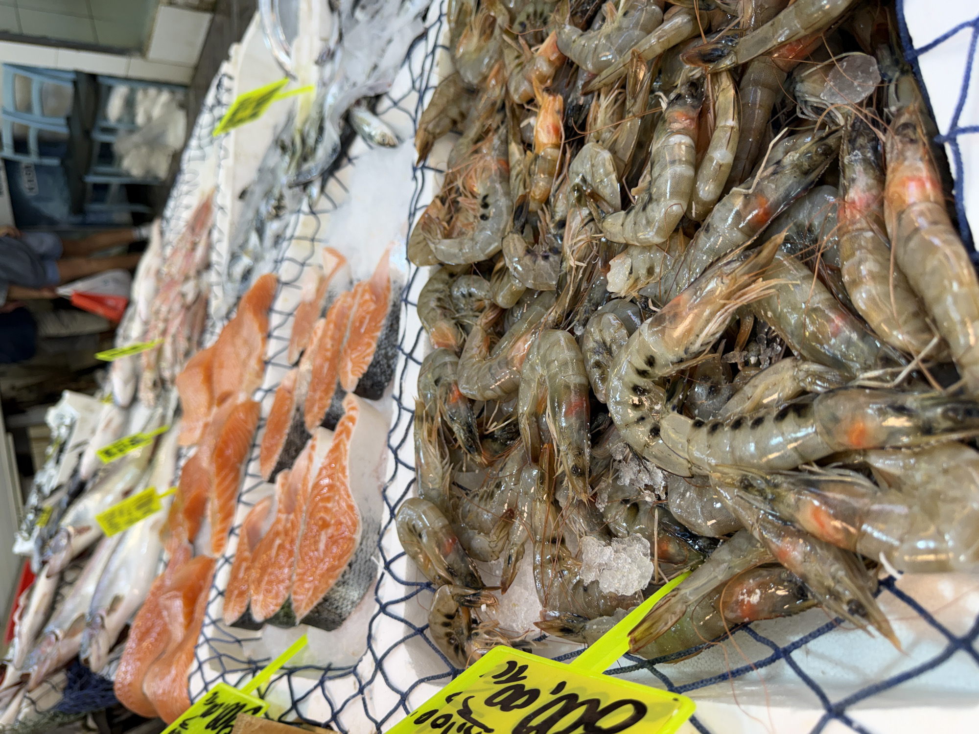 A close-up, eye-level shot captures a seafood stall in Kadıköy, Turkey. In the foreground, a large pile of raw, uncooked shrimp rests on a bed of crushed ice. The shrimp are translucent with some showing hints of orange and red on their bodies. Behind the shrimp, several slices of salmon are arranged on ice. These salmon slices are a vibrant orange-pink color with visible skin on the bottom. Further back, a variety of whole fish, some with scales and fins visible, are displayed. The stall appears to be outdoors or in a covered market, with a glimpse of a person in the background wearing a gray shirt and blue shorts, carrying a red and white bag. Yellow price tags are visible throughout the display, indicating the items for sale.